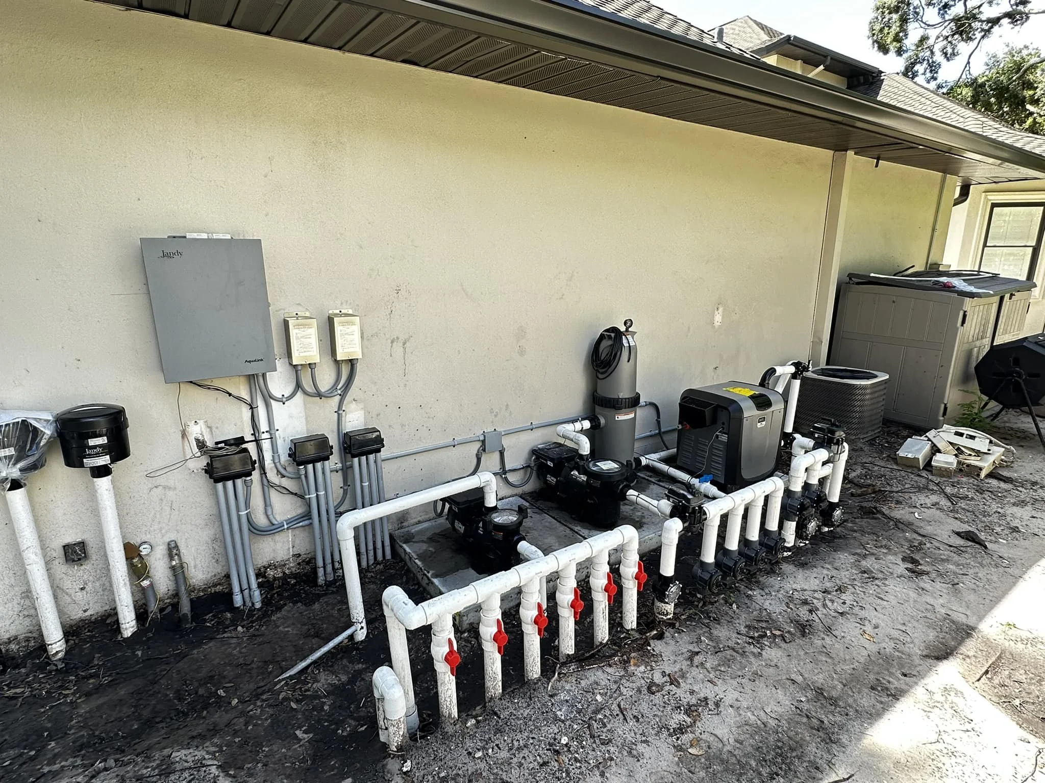 Outdoor view of a pool equipment setup with various pumps, filters, and pipes connected against a beige wall, with a storage box and some miscellaneous items nearby.