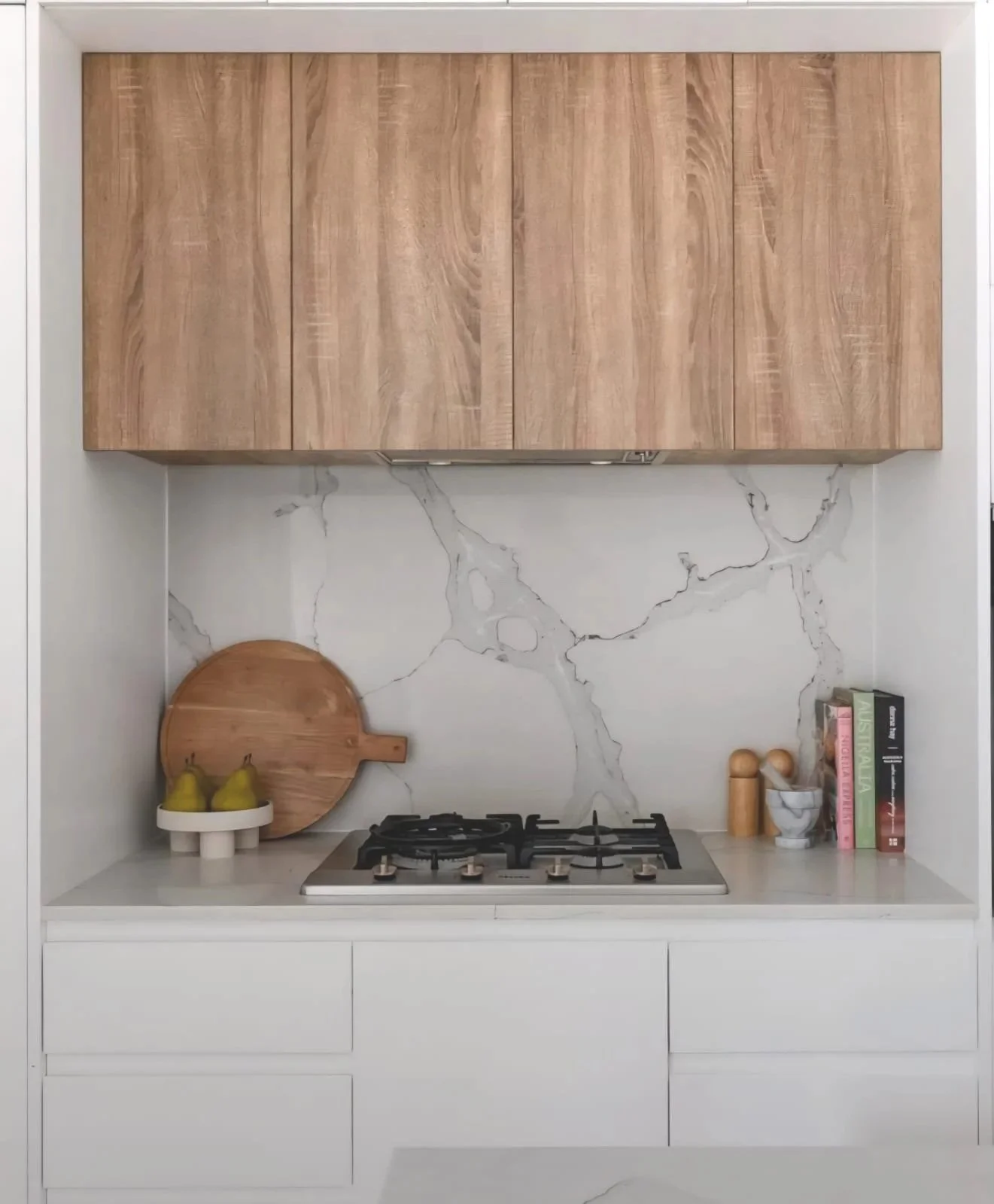 Kitchen with white countertops, wooden cabinetry, marble backsplash, gas stove, decorative pears on a white stand, wooden circular cutting board, salt and pepper shakers, a marble cup, and books.