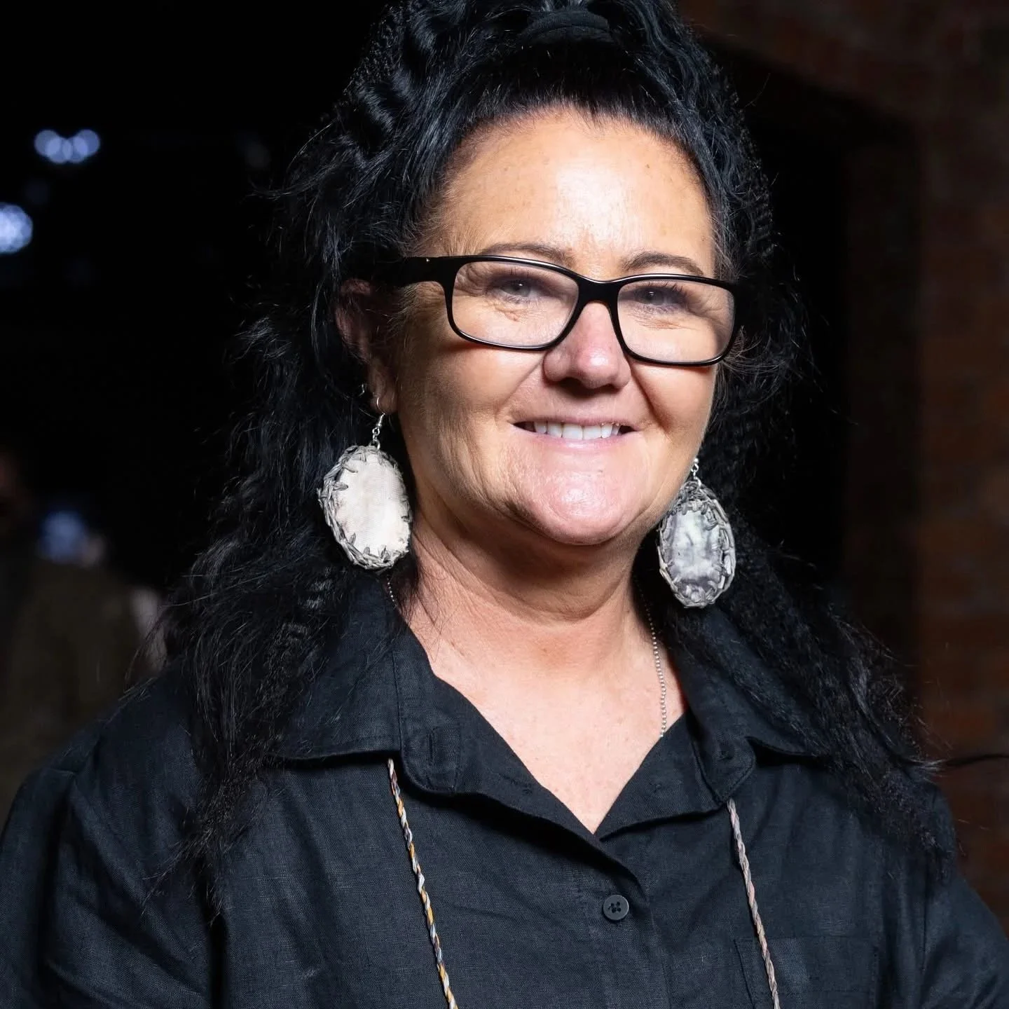 An indigenous woman with black hair, wearing glasses and large earrings, smiling and dressed in black.
