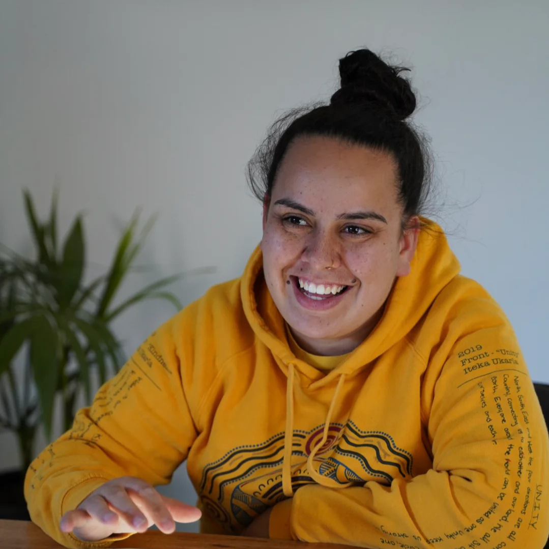 A young indigenous woman, who specialises in creating digital art, with dark hair tied in a bun, smiling, wearing a yellow hoodie with black designs and text on the sleeves, sitting at a table.