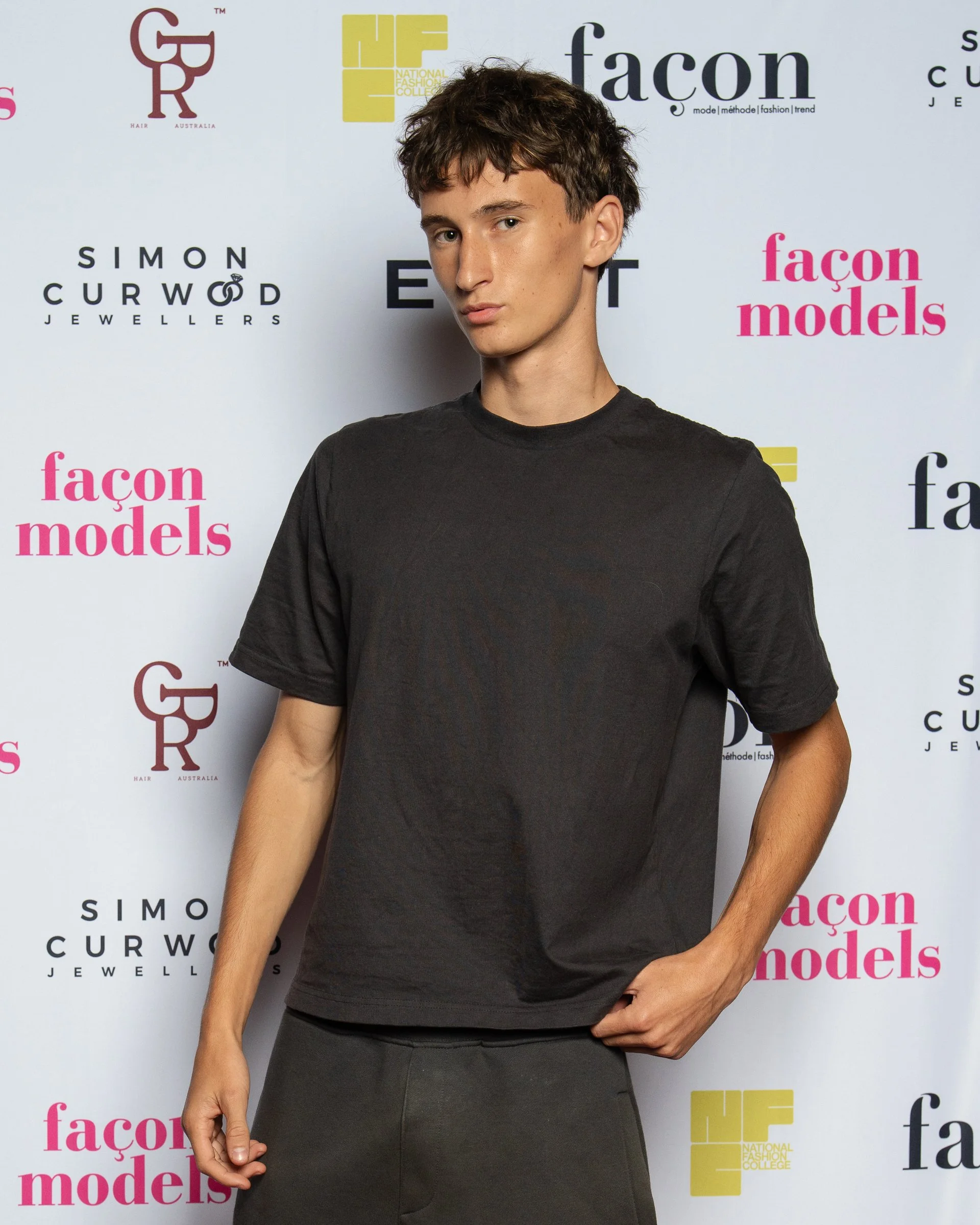 A young woman with short brown hair, wearing a black t-shirt, standing in front of a white backdrop with logos of 'façon models,' 'Simon Curwood Jewellers,' and 'National Fashion College.'