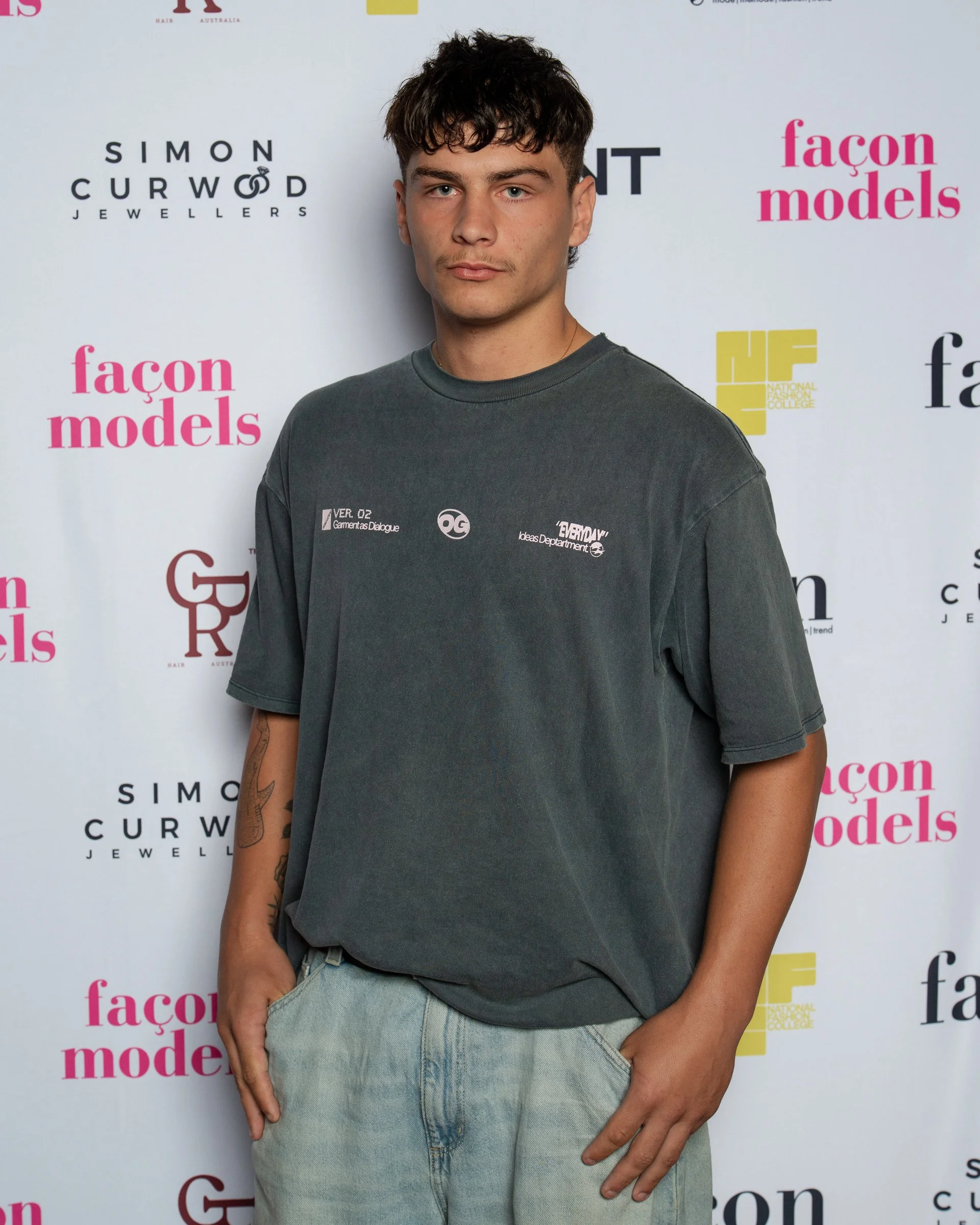 A young man with brown hair, light skin, and blue eyes standing in front of a step and repeat backdrop with logos, wearing a dark gray T-shirt and light-colored jeans.