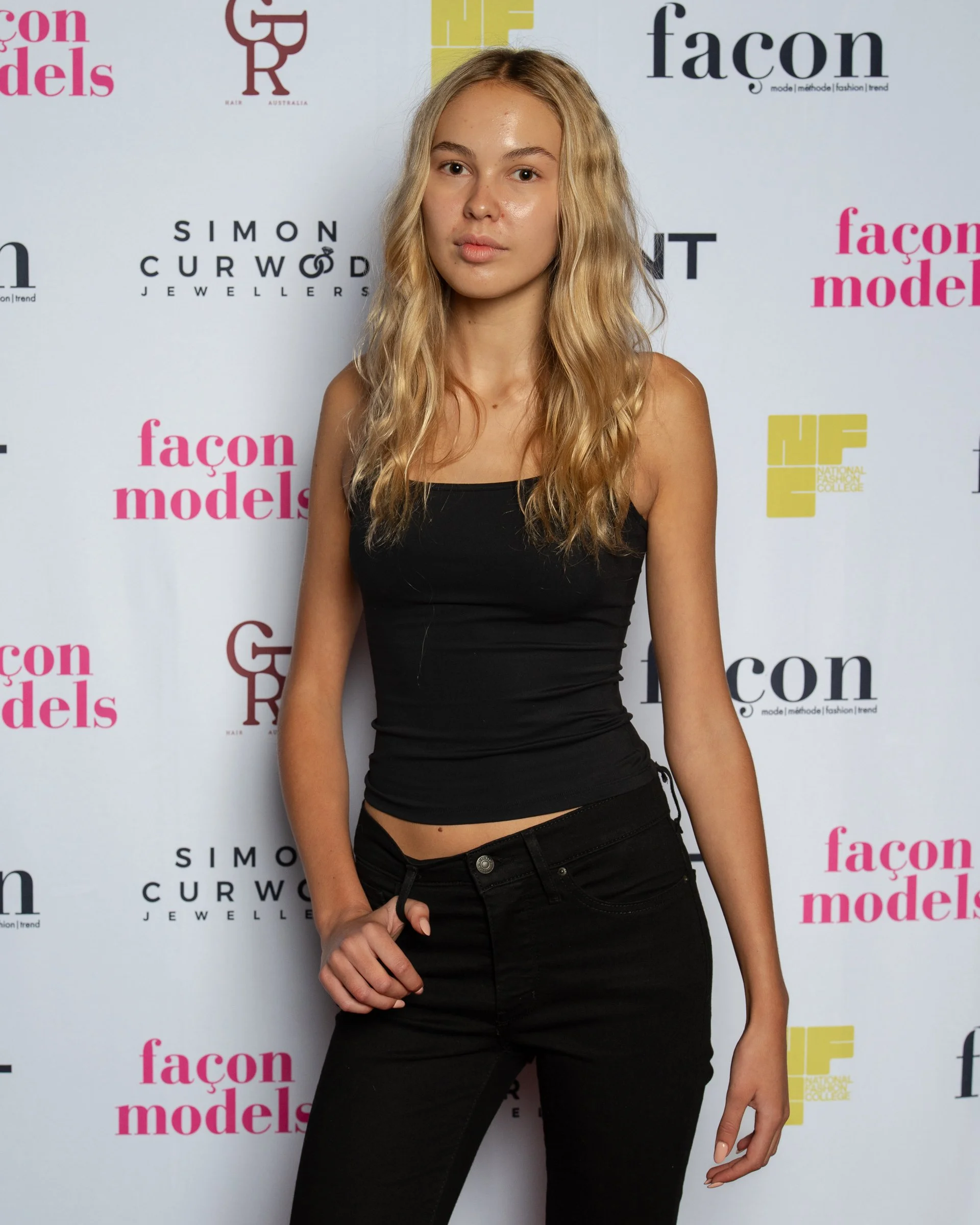 A young woman with long, blonde wavy hair wearing a black tank top and black pants standing in front of a white backdrop with pink and black logos for fashion and modeling organizations.