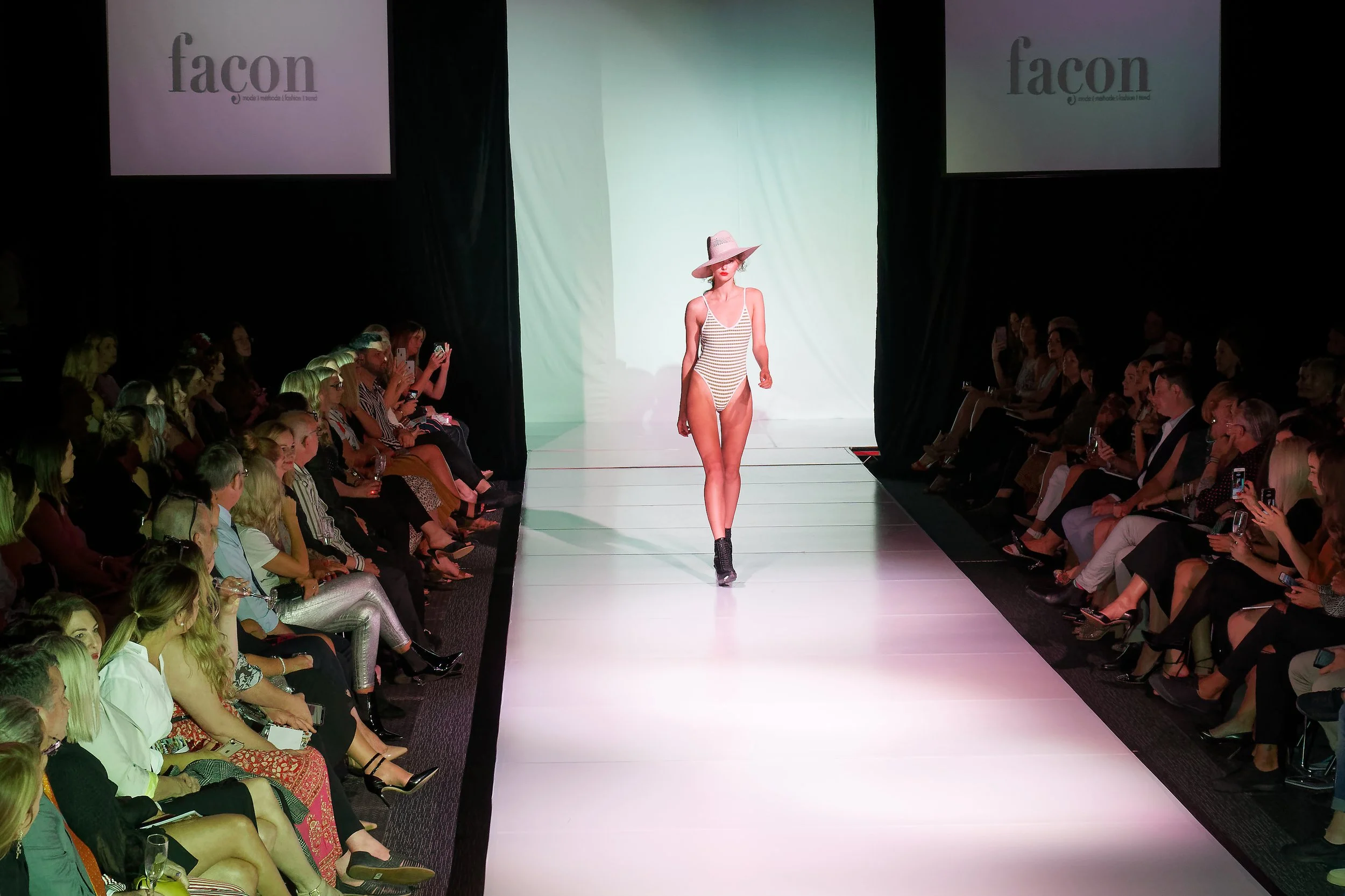 A fashion runway show with a model walking down the runway wearing a striped swimsuit and a wide-brimmed pink hat. The audience is seated on either side of the runway, watching and taking photos. The background has two large screens displaying the word 'facon' and a tagline beneath.