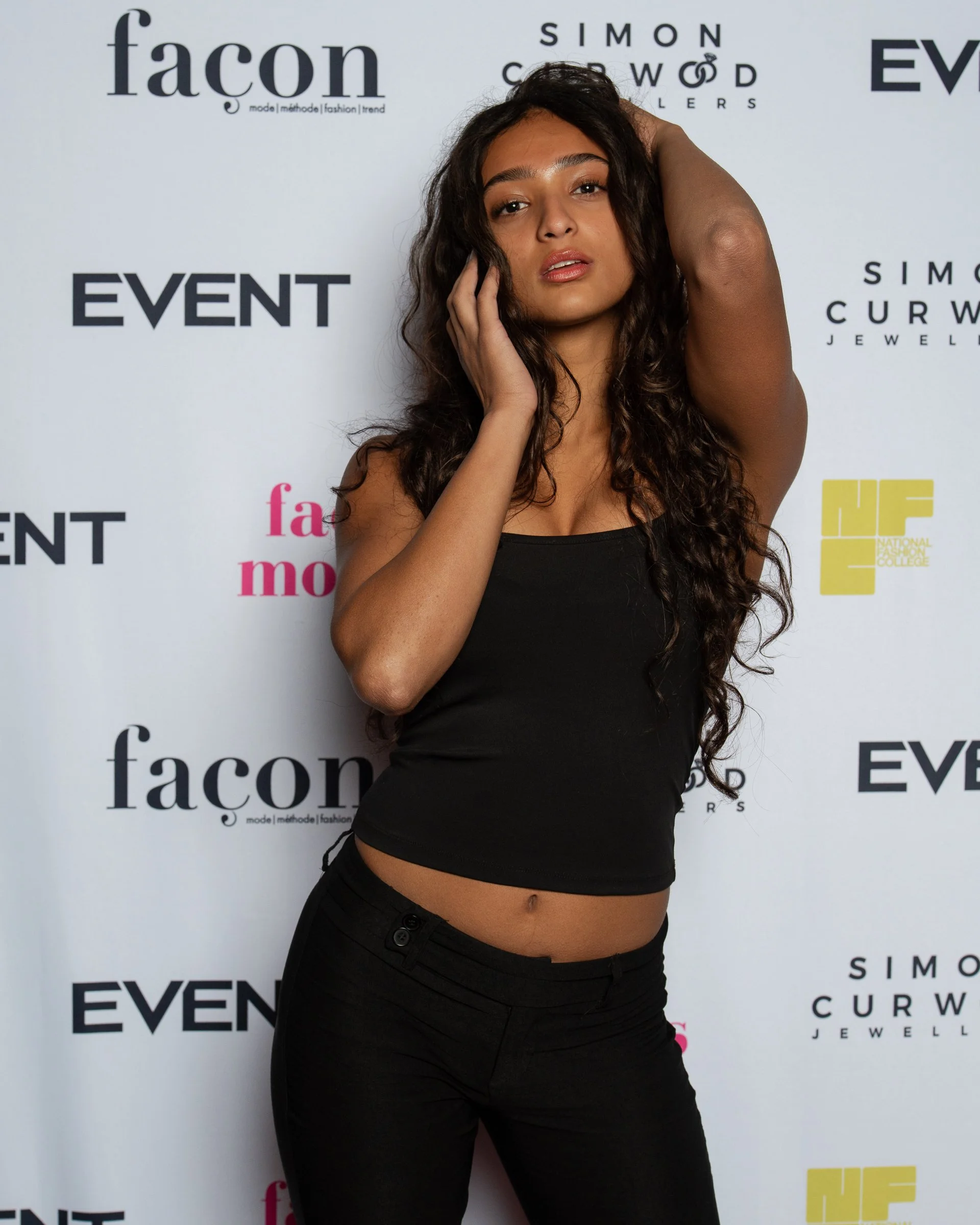 A woman with long curly hair posing with her hand on her head and her other hand touching her face in front of a backdrop with logos and text, including 'fئçon', 'Simon Curwood Jewelry', and 'EVENT'.
