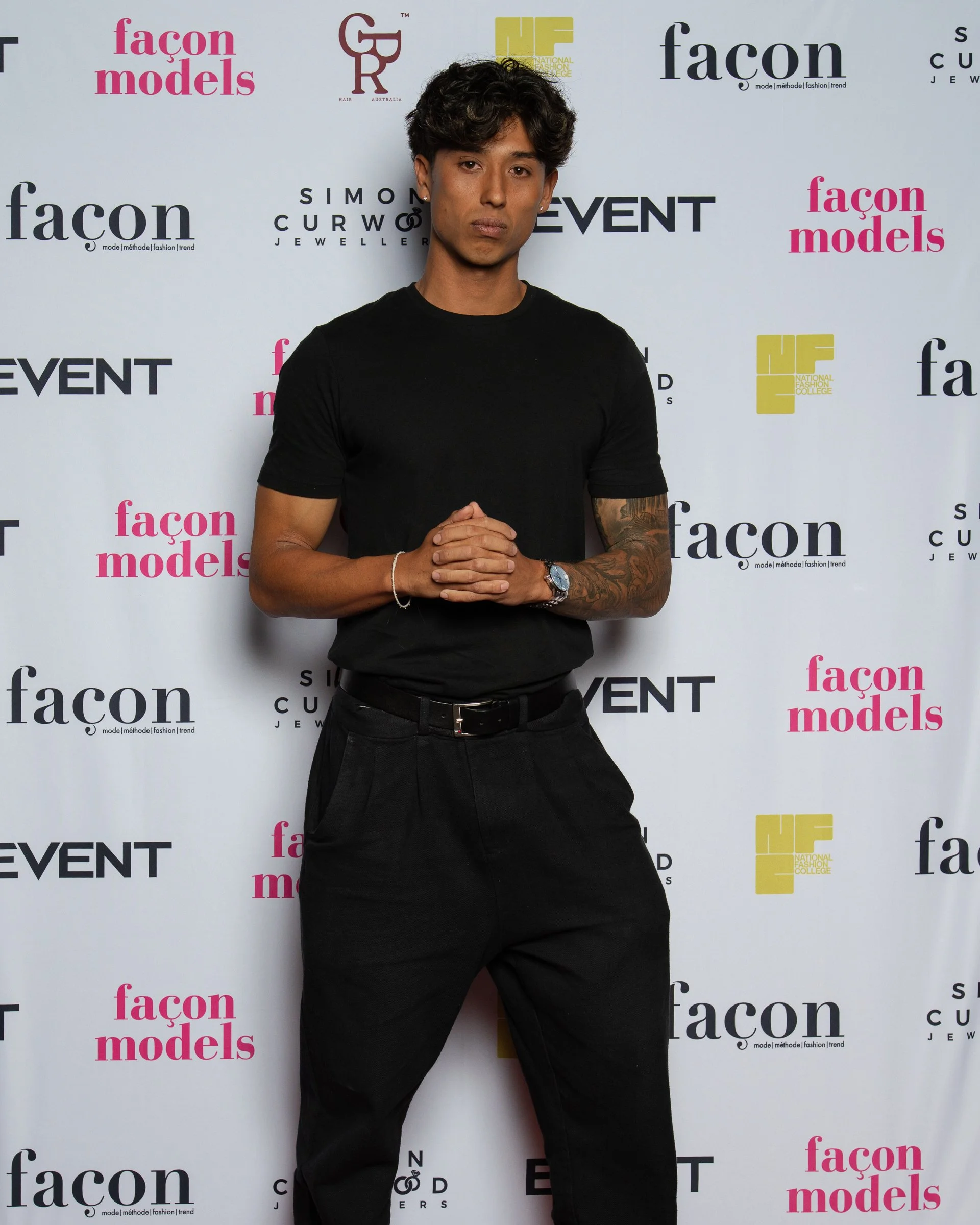 Young man with dark wavy hair, wearing a black t-shirt and black pants, standing against a step and repeat backdrop with logos from 'Façon Models,' 'Simon Curwel Jewelry,' and others, at an event.