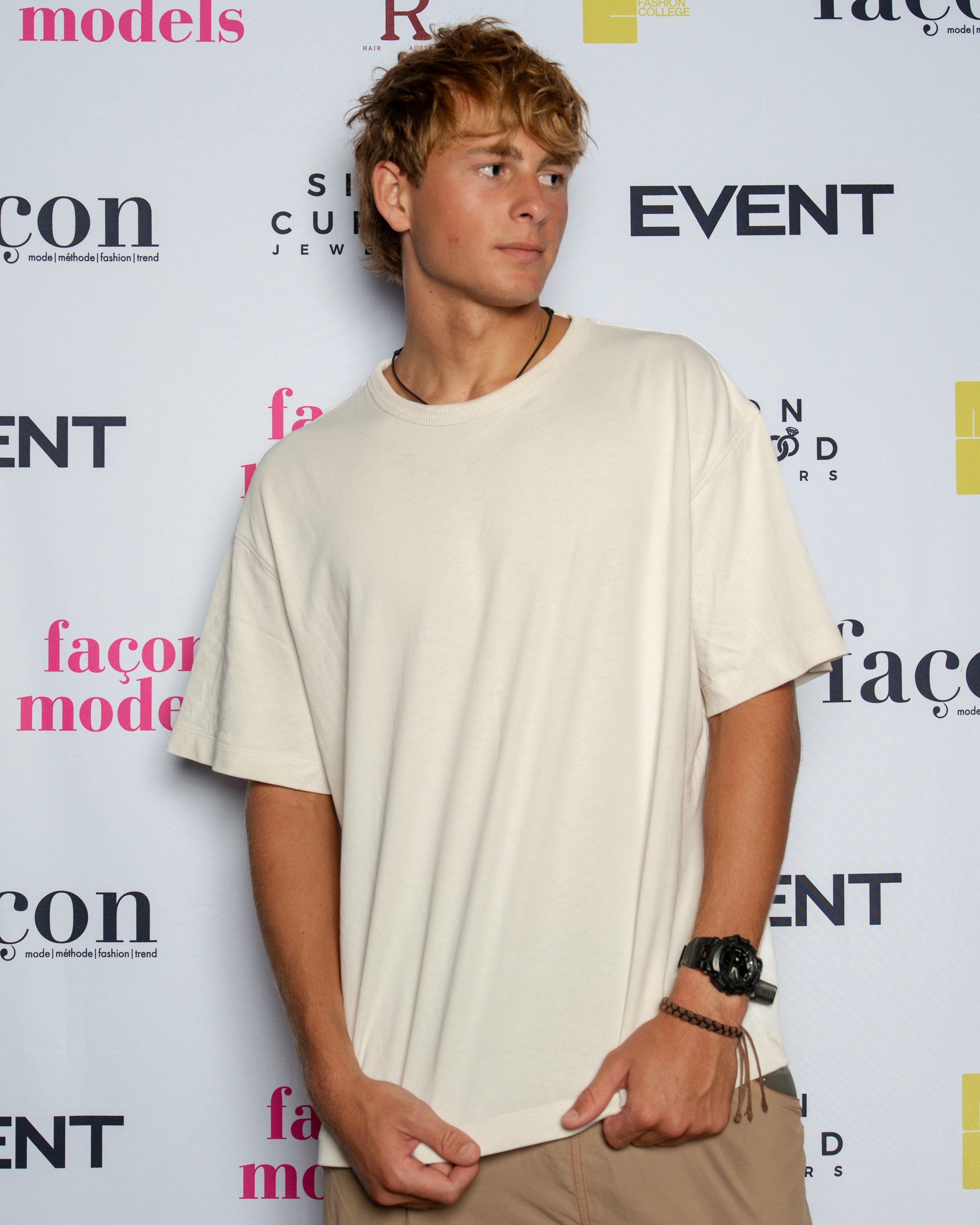 A young man with blonde hair, wearing a beige oversized t-shirt, beige pants, a black wristwatch, and a brown bracelet, is standing in front of a backdrop with various magazine and fashion event logos, looking to his right with a neutral expression.