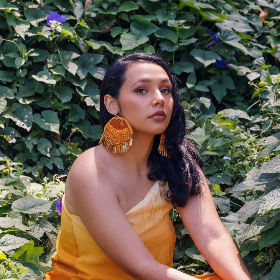 An indigenous woman with dark hair, posing for a fashion magazine, sitting infront of a backdrop of green leaves, wearing a bright yellow dress and matching bold earrings.