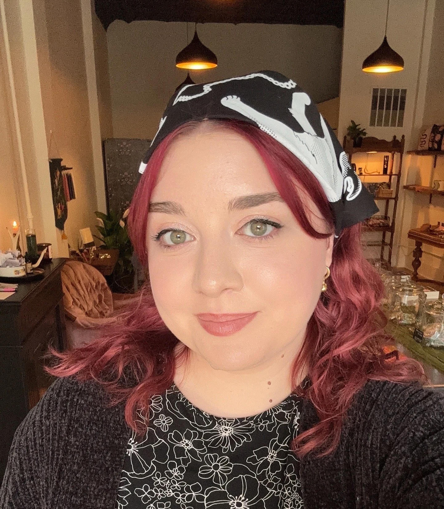 A femme-presenting person with red wavy hair wearing a black and white bandana and black top with white floral pattern, in a warmly lit room with shelves, furniture, and decor.