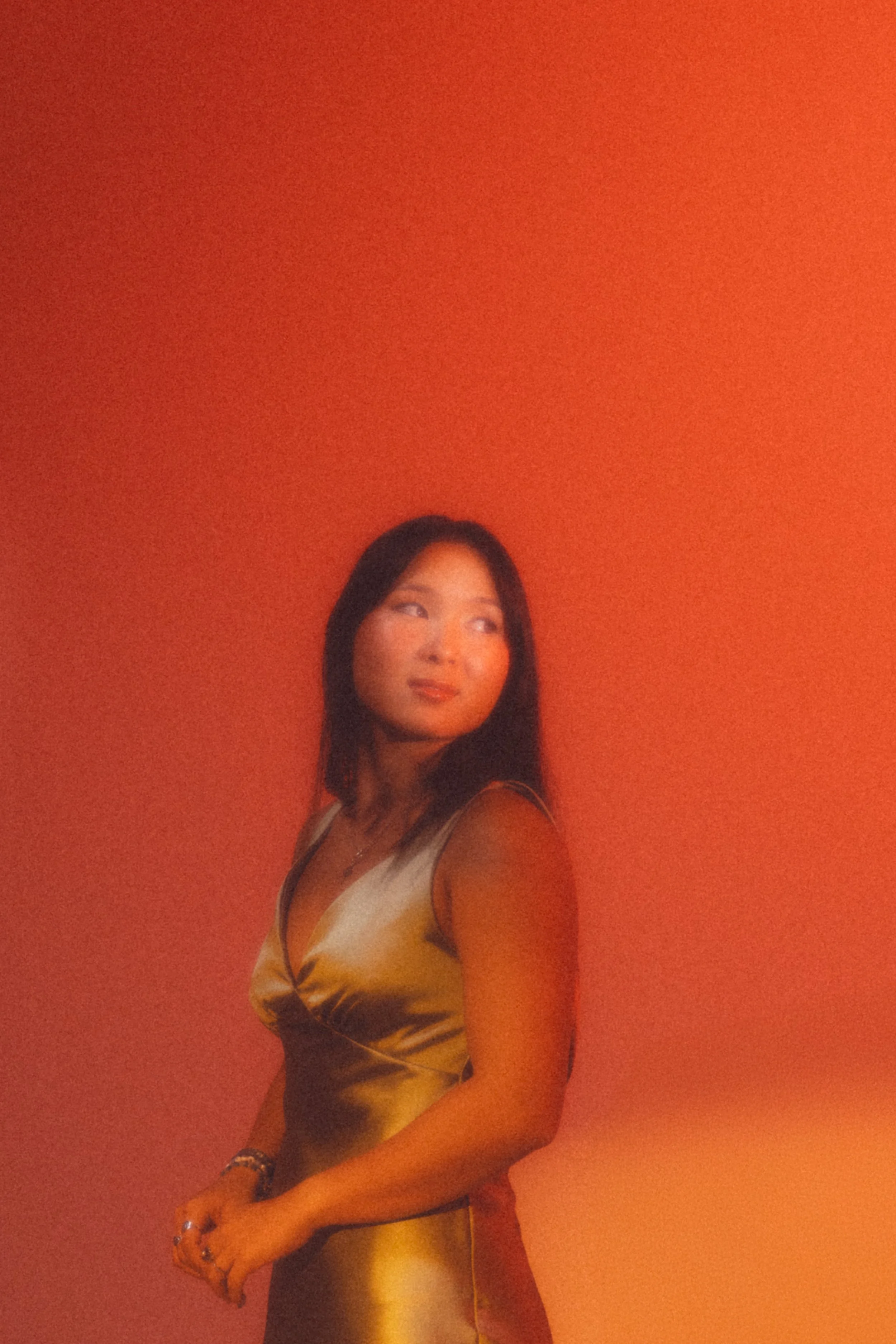 A woman with long dark hair standing against an orange wall, wearing a shiny gold satin dress, looking to her left with her arms clasped in front of her.