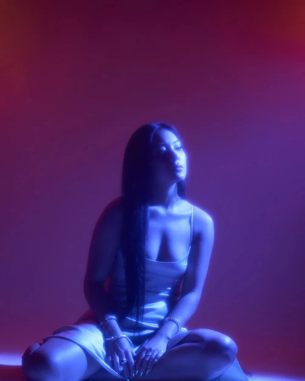 Woman sitting cross-legged in a colorful, moody lighting setting with purple and pink hues, wearing a light dress with thin straps.