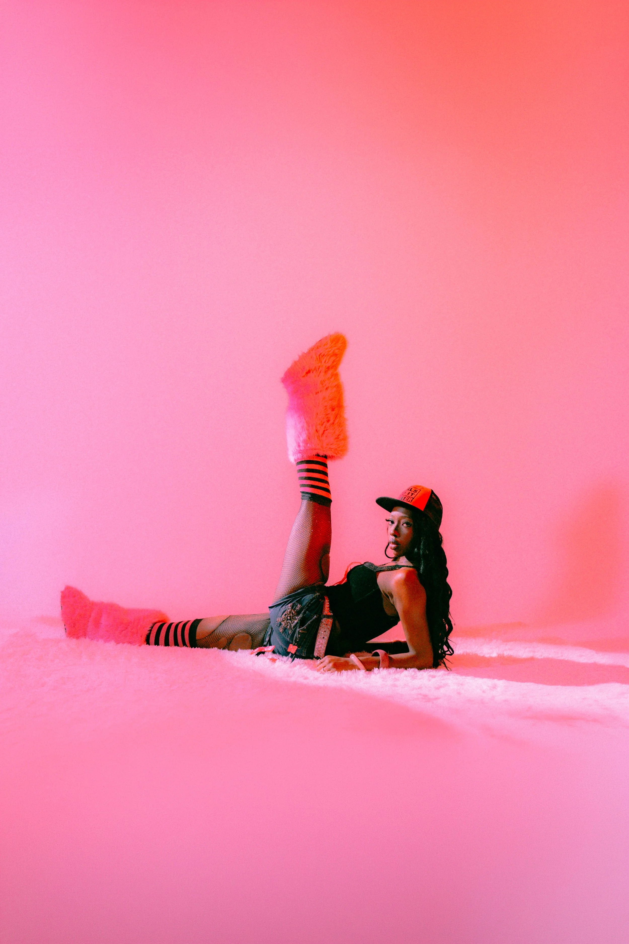A woman with long black hair wearing a black top, shorts, fishnet stockings, striped leg warmers, and a baseball cap poses on a pink fluffy carpet with a matching pink background. She has one leg lifted high and is lying on her side, supporting herse