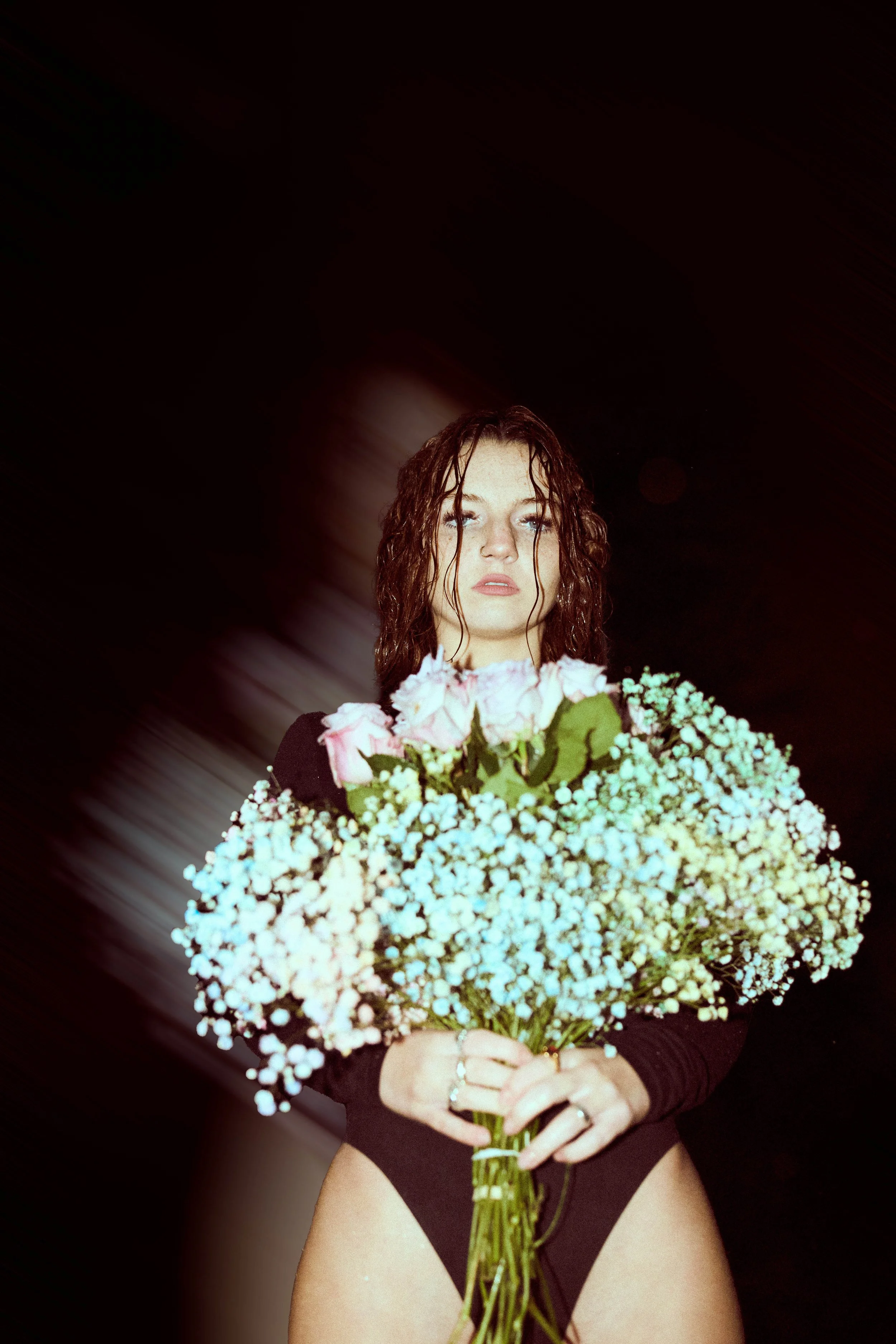 A woman with wet hair holding a bouquet of pink and white flowers, dressed in black lingerie, against a dark background.