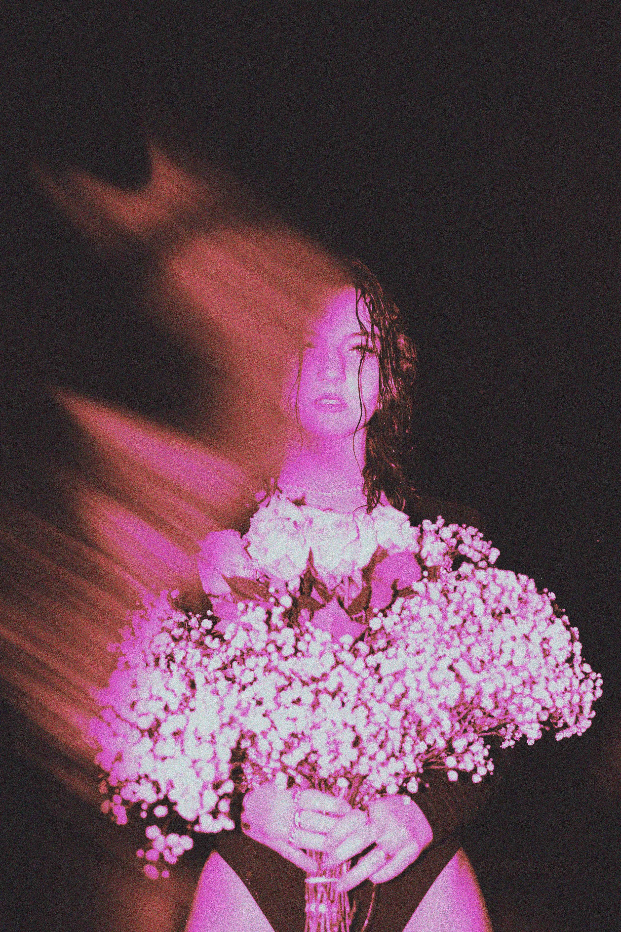 A woman with curly hair holding a large bouquet of pink and white flowers, wearing underwear, against a dark background with pink lighting effects.