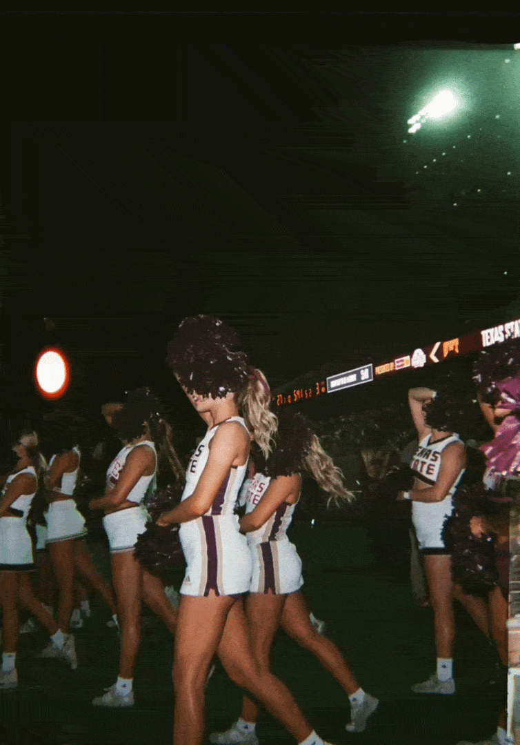 Cheerleaders in white uniforms with purple and gold accents perform during a sports event at an indoor stadium.
