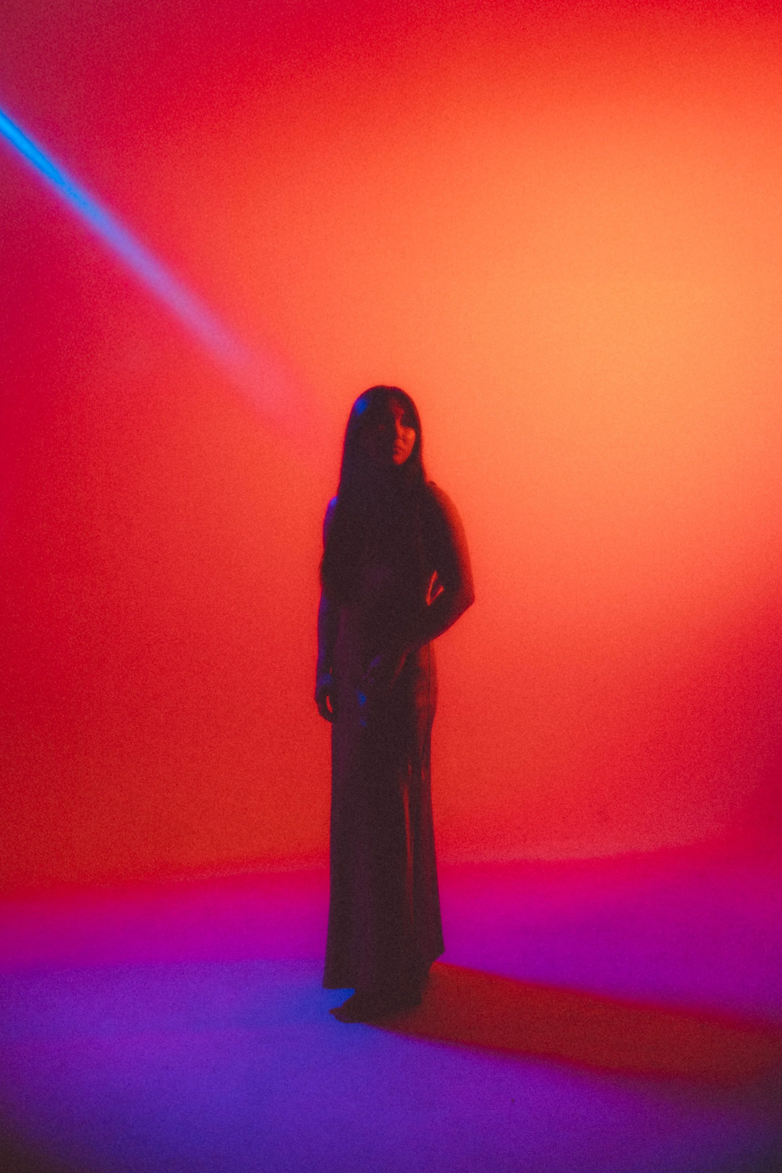 Person standing against a colorful, gradient background with shades of red, orange, pink, purple, and blue, silhouetted and facing forward.