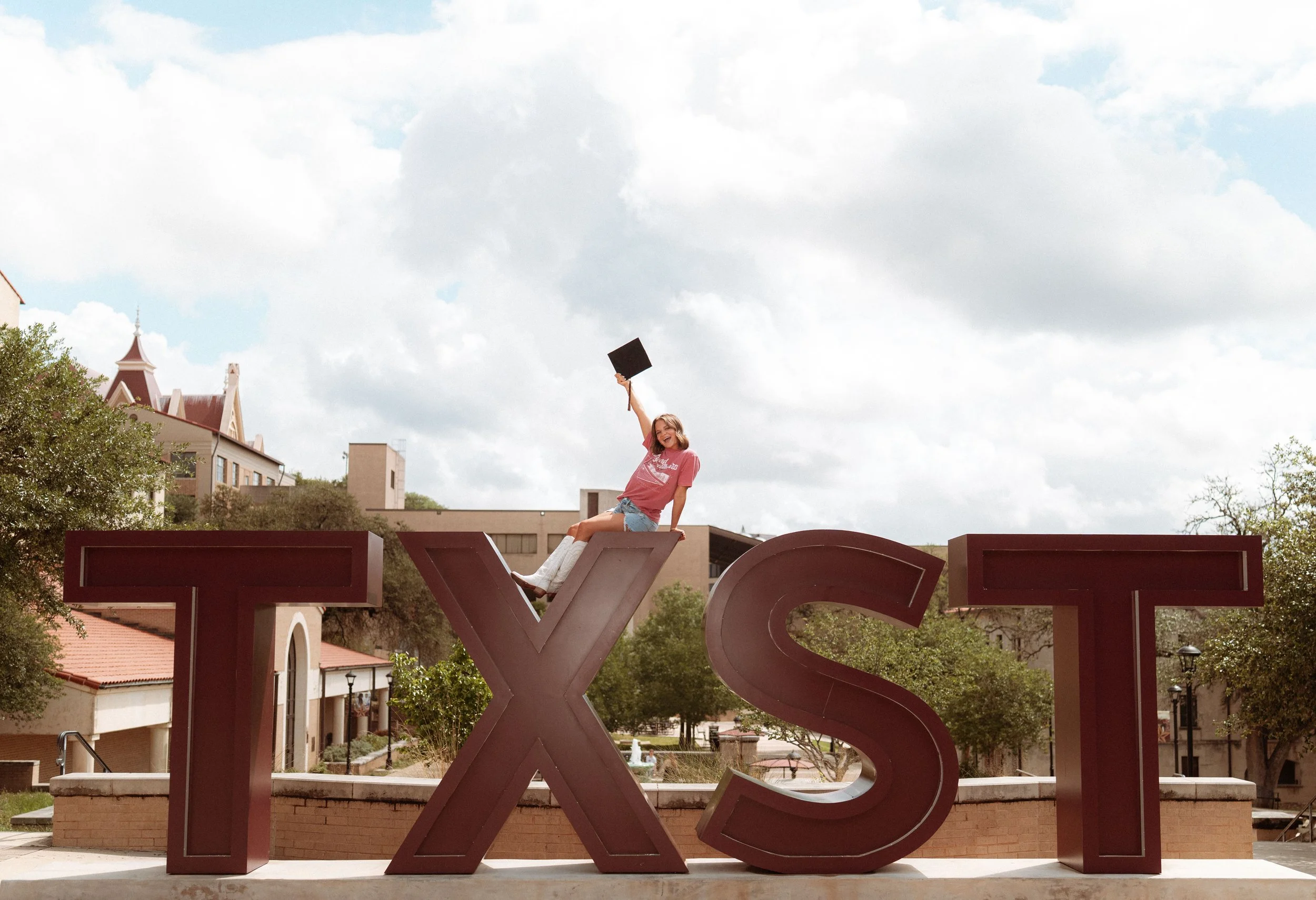 A young woman sitting on top of a large red letters spelling 'LOVE,' raising a black square object in the air, outdoors on a cloudy day with buildings and trees in the background.