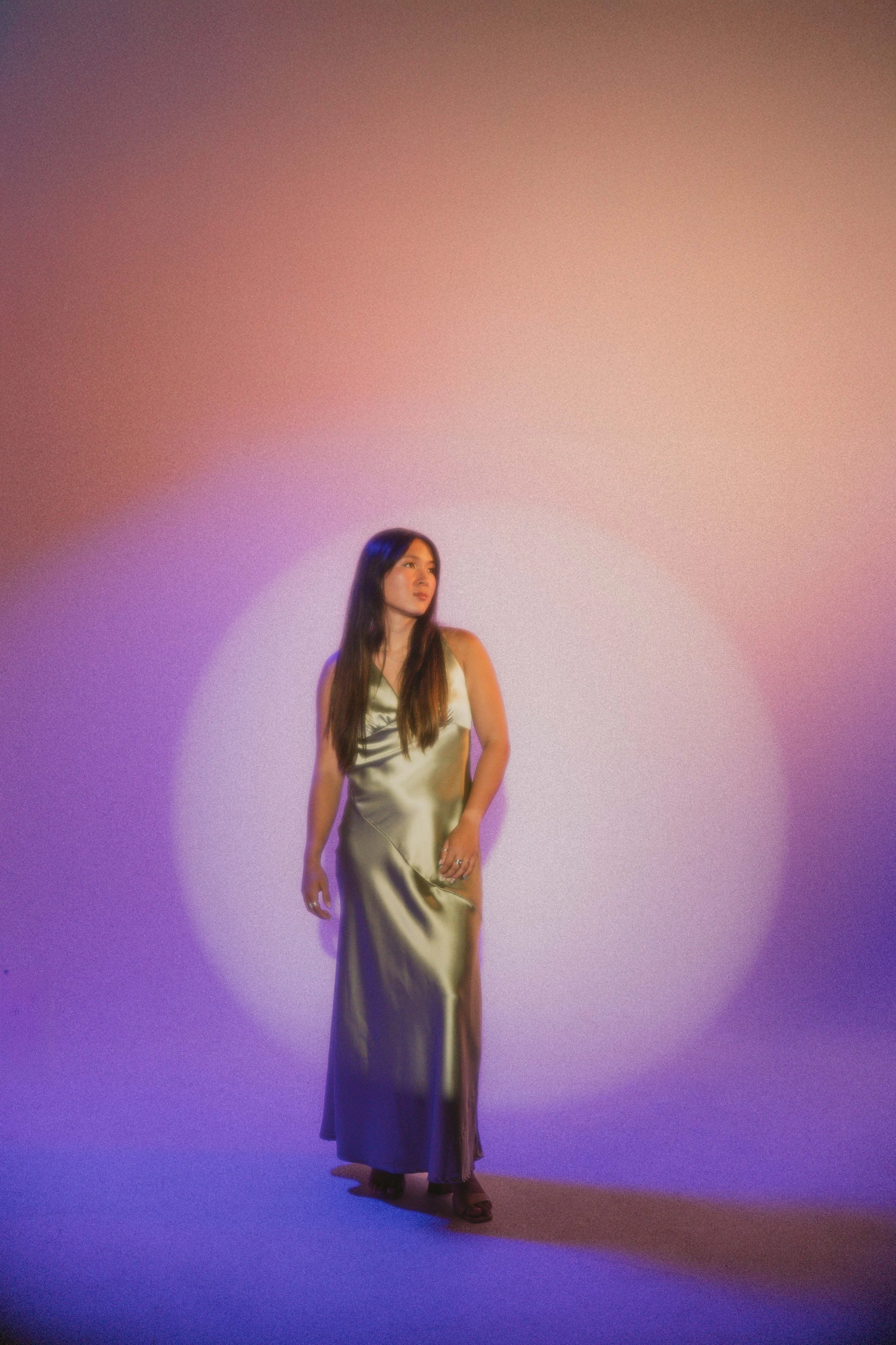 A woman with long dark hair wearing a satin silver evening gown and high heels standing against a colorful gradient background.