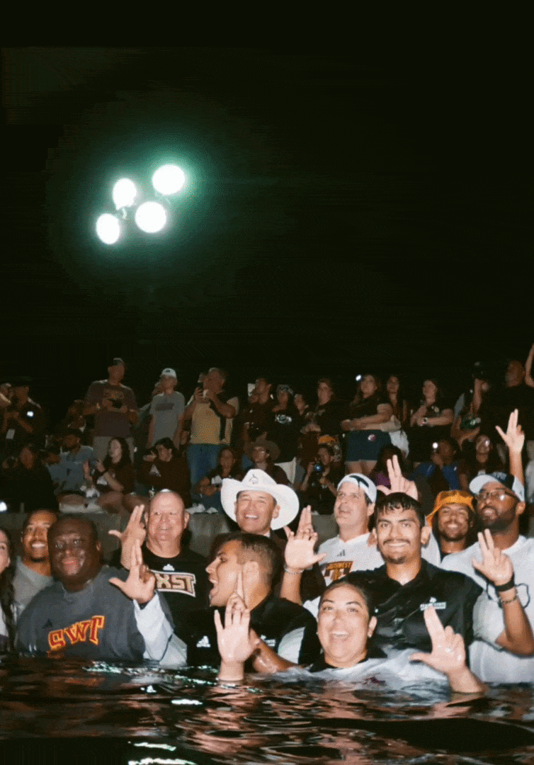 Group of people celebrating in a pool at night, with some making peace signs, wearing casual clothes and hats, while others stand in the background under bright lights.