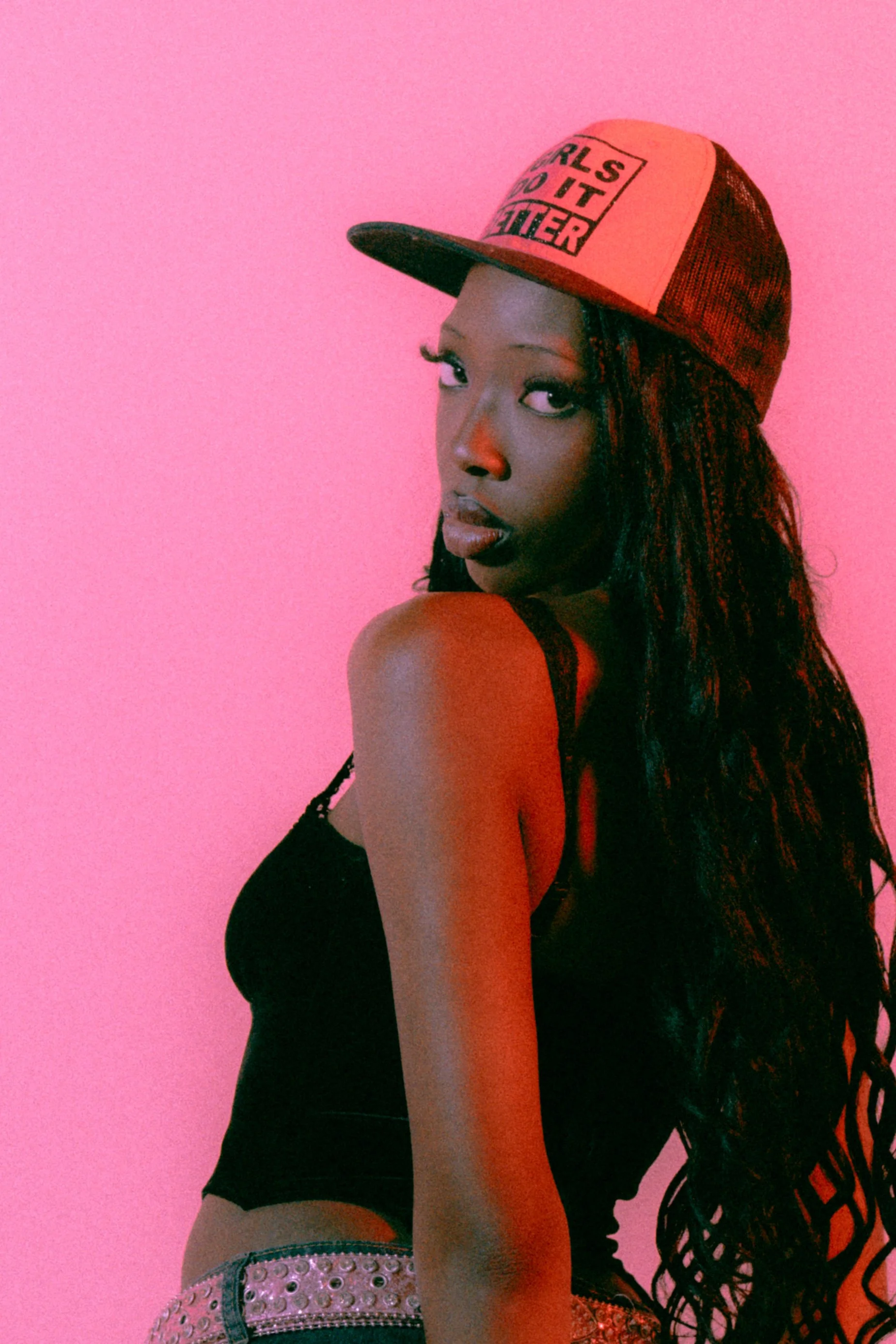 A woman with dark skin and long curly hair wearing a colorful cap and a black tank top, posing against a pink background.