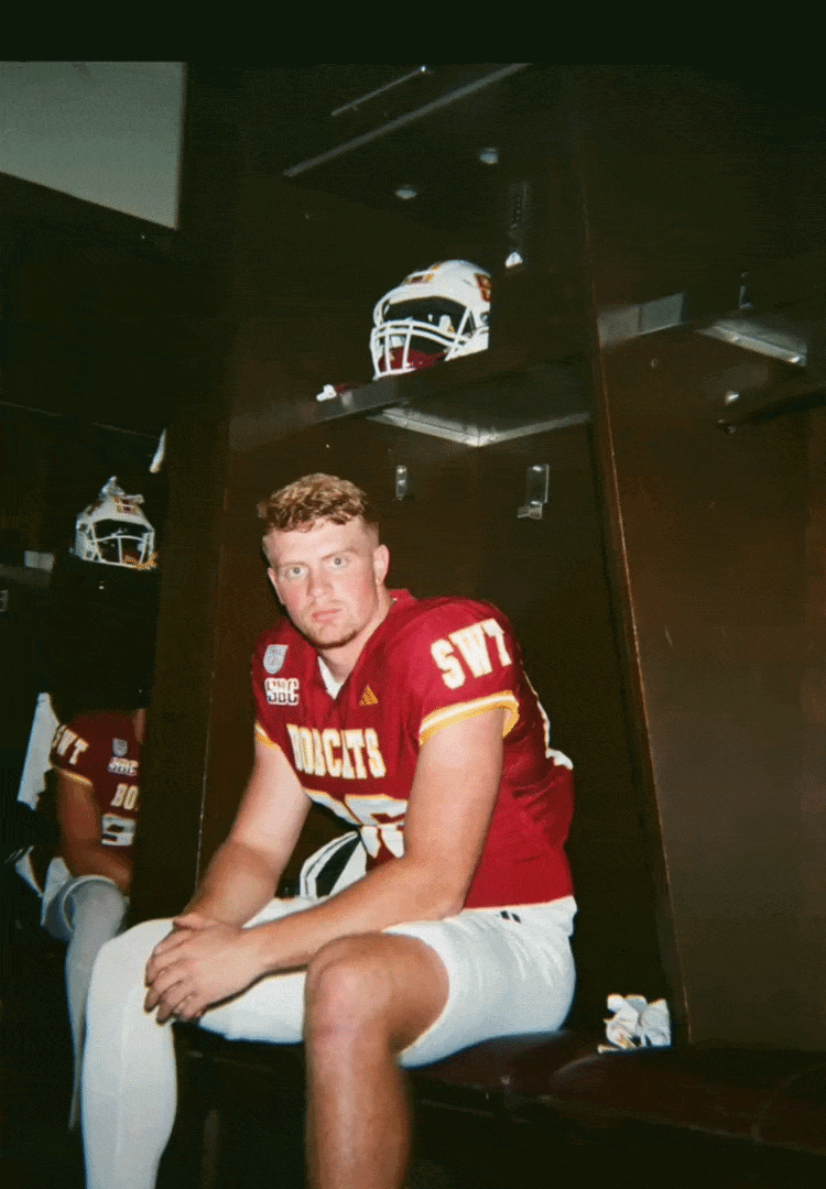 A football player sitting in a locker room wearing red and white uniform with the number 99, and a helmet placed on the top shelf of the locker behind him.
