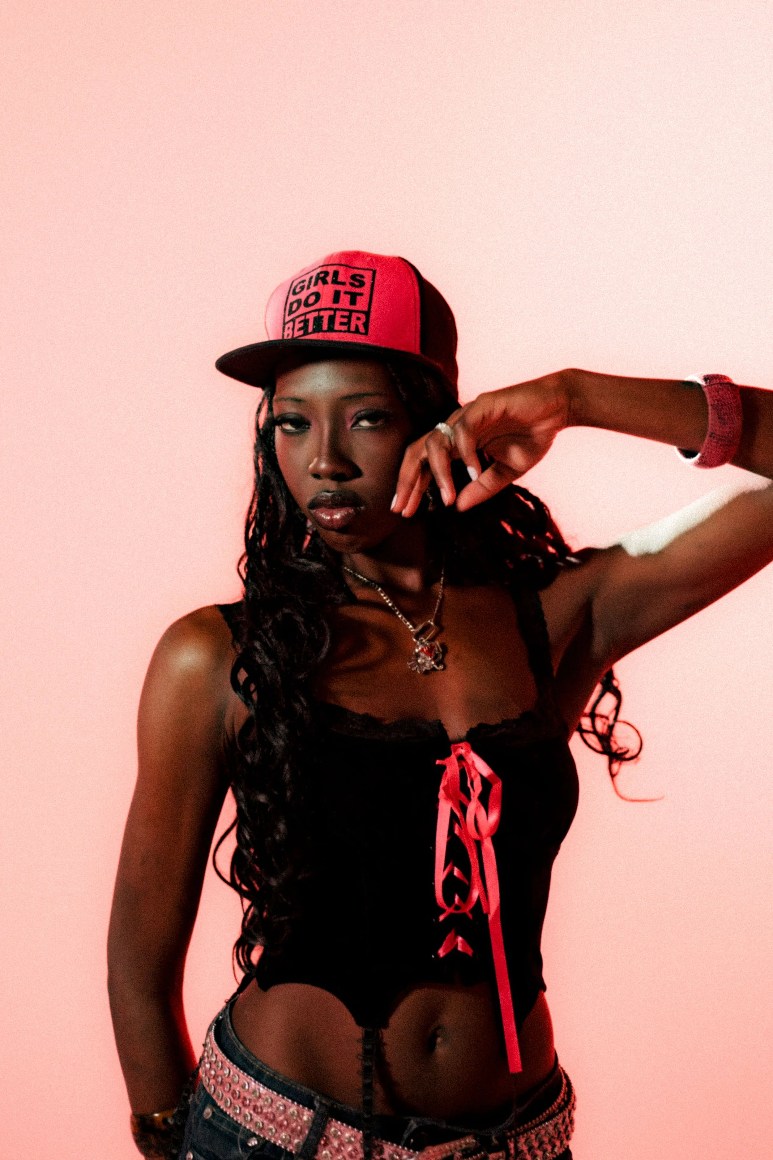 A woman with long, curly hair wearing a red and black cap that says "Girls Do It Better". She has dark lipstick and is dressed in a black outfit with a choker necklace, denim shorts with pink rhinestones, and a red ribbon detail. She is posing with h