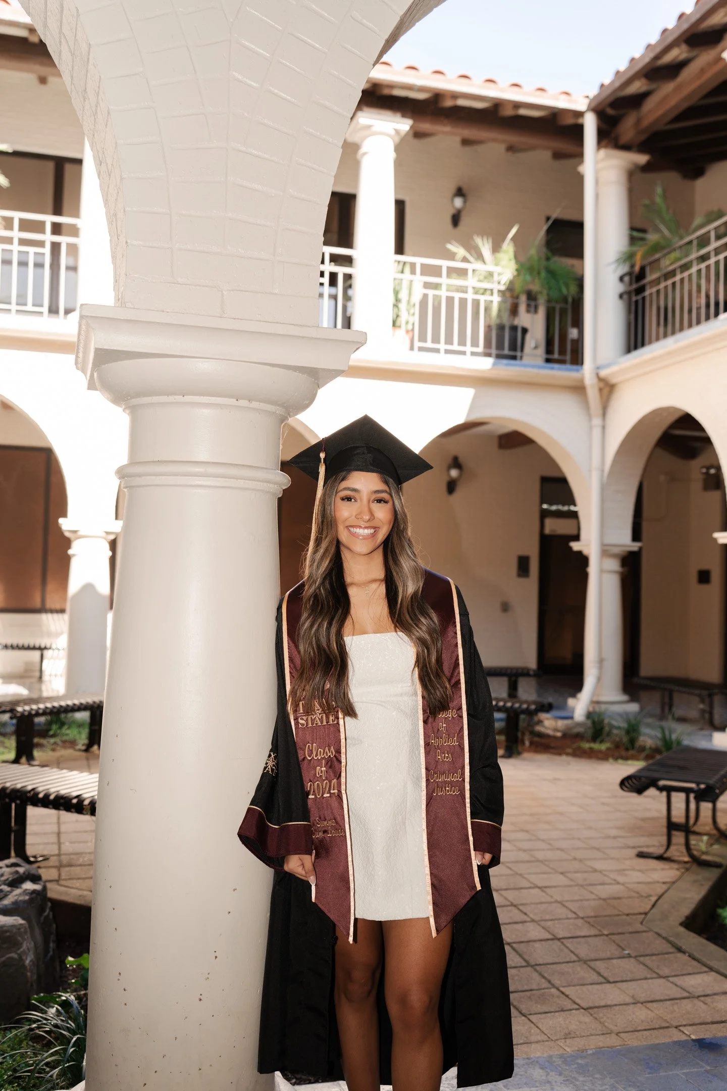 A woman in a graduation cap and gown stands outdoors in front of an architectural courtyard, smiling at the camera. She is wearing a white dress and a maroon and gold stole that reads 'Class of 2024' from California State University.
