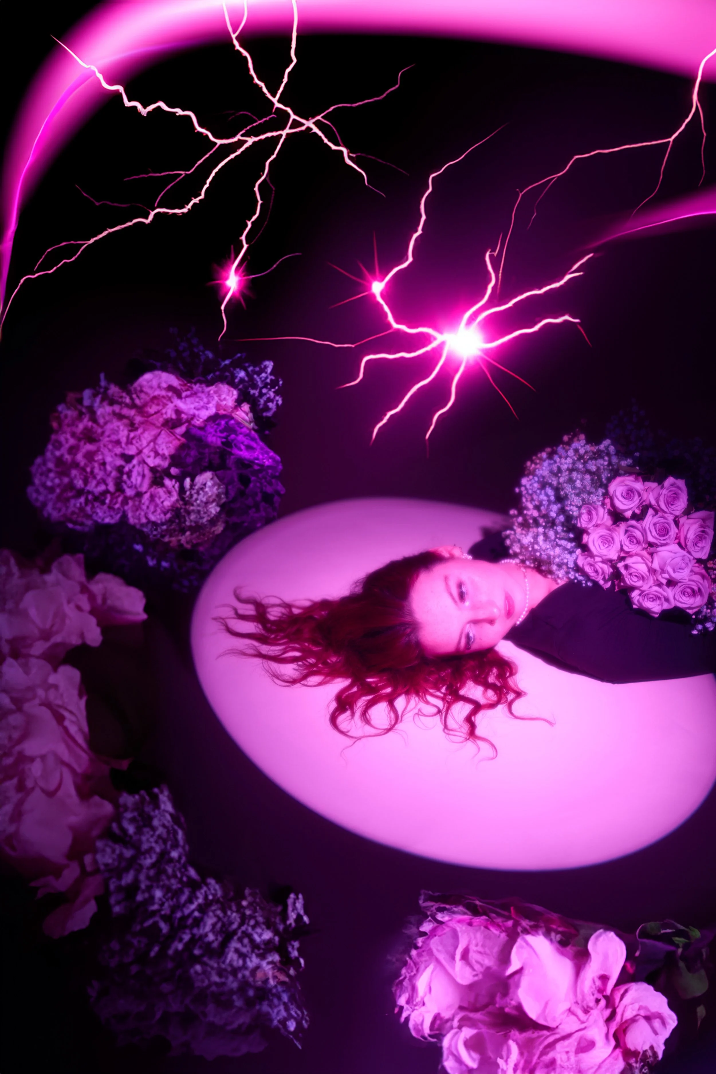 A woman with long red hair lying in a pink-lit bathtub surrounded by pink and purple flowers and purple lightning effects in the background.