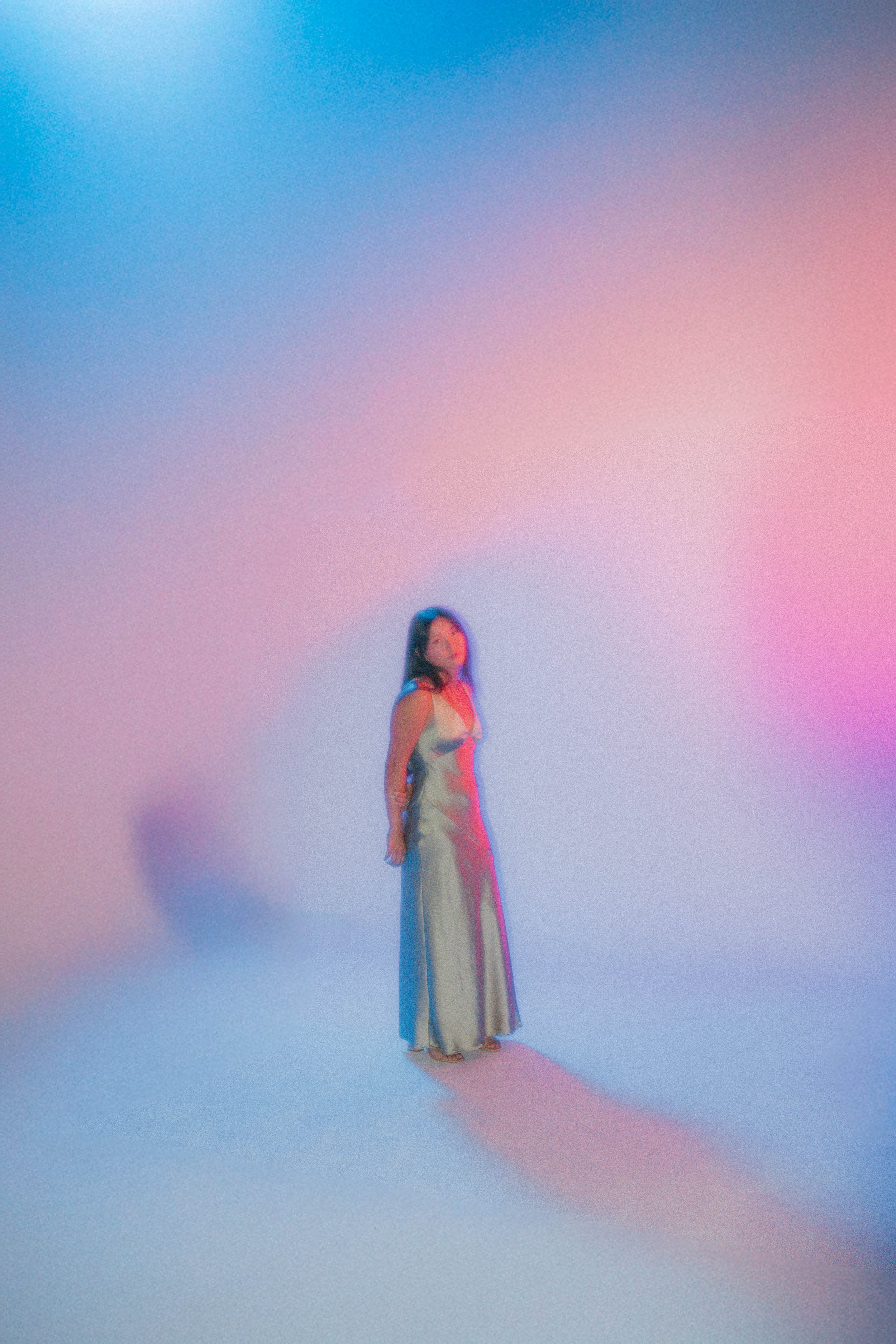 A woman in a long white dress stands on a pastel-colored gradient background with soft pink and blue hues. She is positioned towards the center, looking slightly to the side, with her hands at her sides.