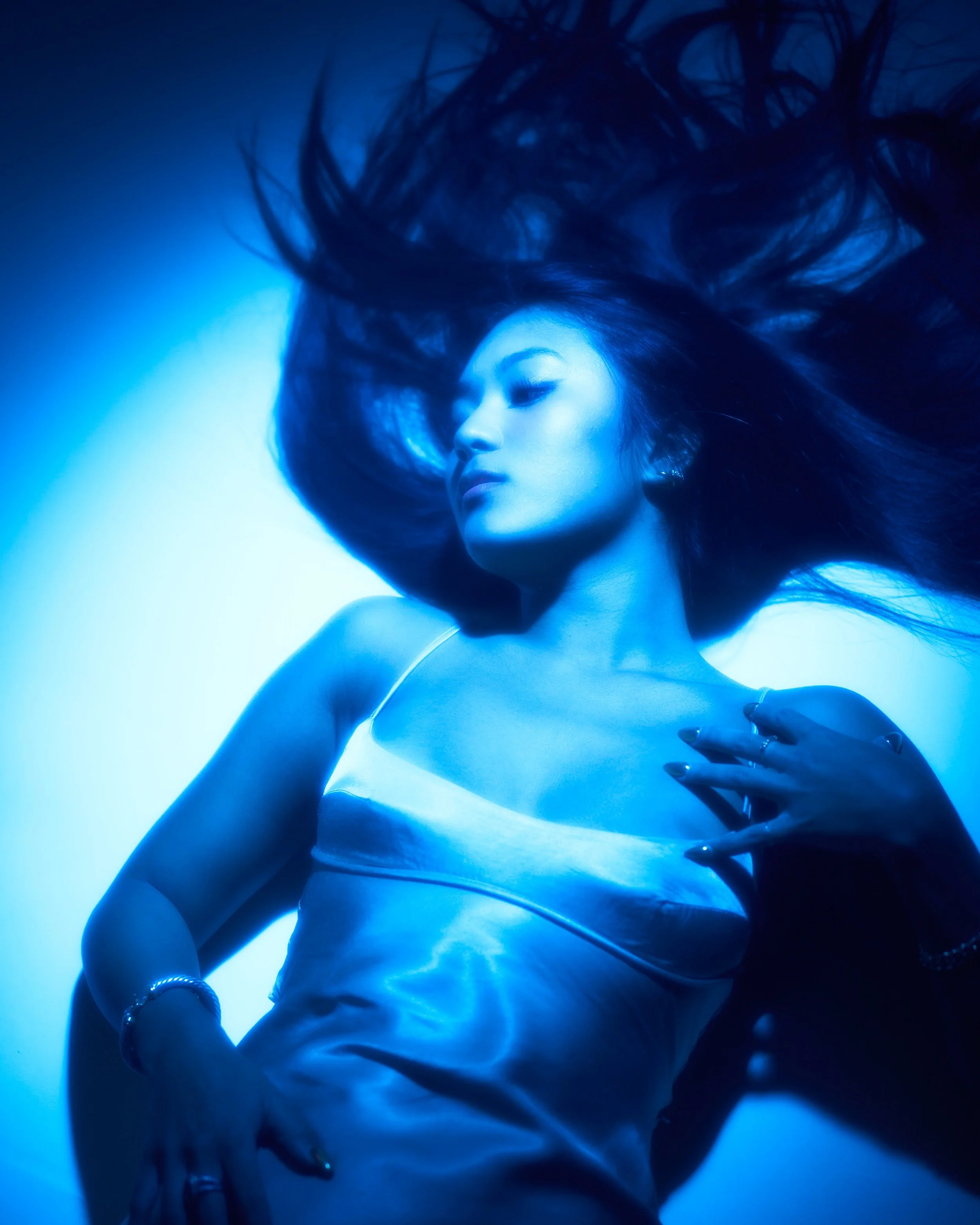 A woman with long dark hair lying down, illuminated with blue lighting, wearing a white tank top and jewelry, with her eyes closed and hand resting on her chest.