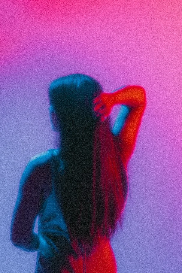 Silhouette of a woman with long hair, standing against a pink and purple gradient background, with one arm raised behind her head.