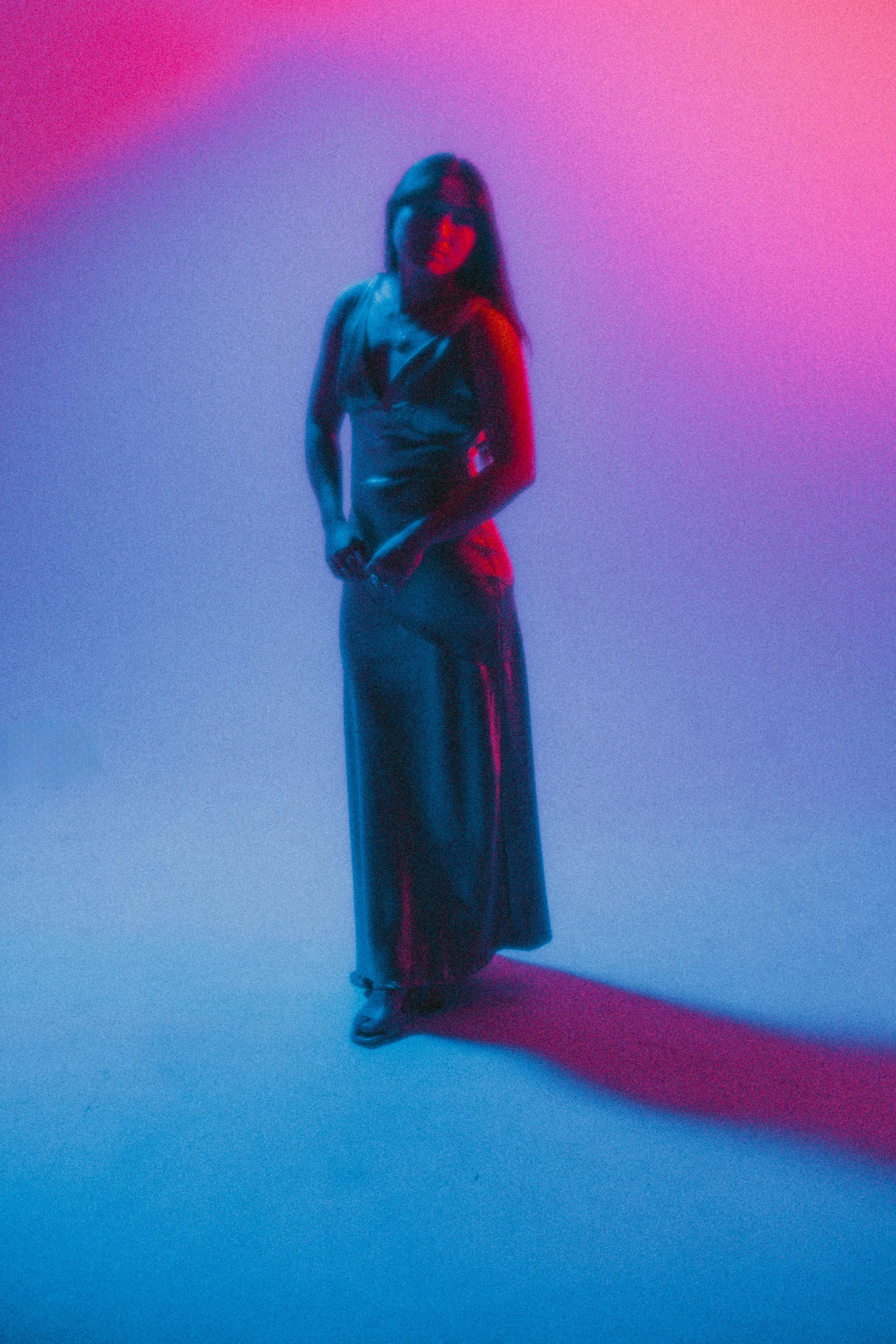 A woman standing on a colorful gradient background with pink and blue lighting, wearing a shiny outfit and sunglasses.
