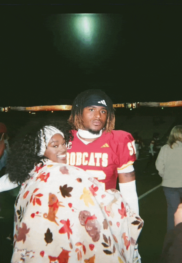 A woman and a young man at a football game, with the woman wrapped in a blanket with autumn leaves and animals, and the man wearing a red football jersey with "BOBCATS" on it, standing under stadium lights.