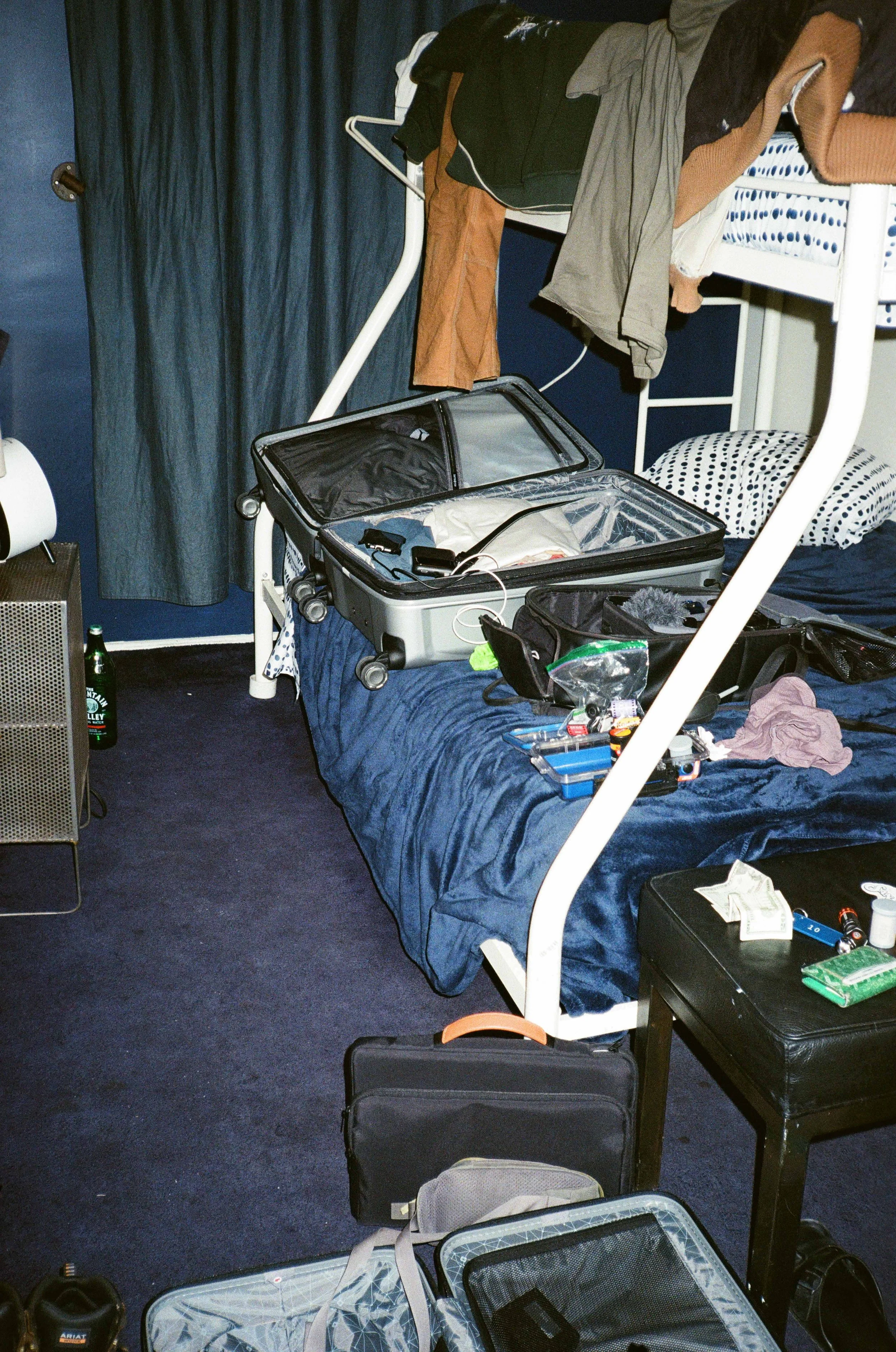 A cluttered hotel room with an unmade bed, open suitcase, various clothing items, and travel essentials scattered around. There are suitcases, a nightstand, a chair, and personal belongings including a water bottle, snacks, and electronic devices.