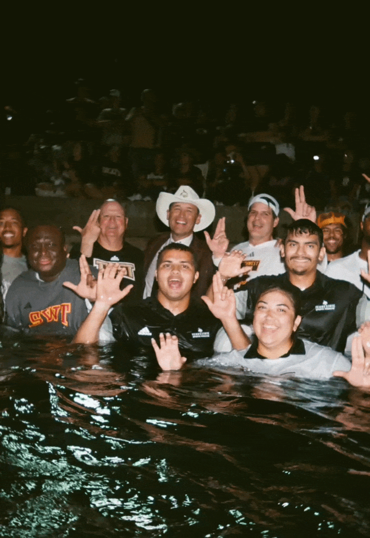 Group of people standing in water at night, smiling and making peace signs, some wearing uniforms and one person in a cowboy hat.