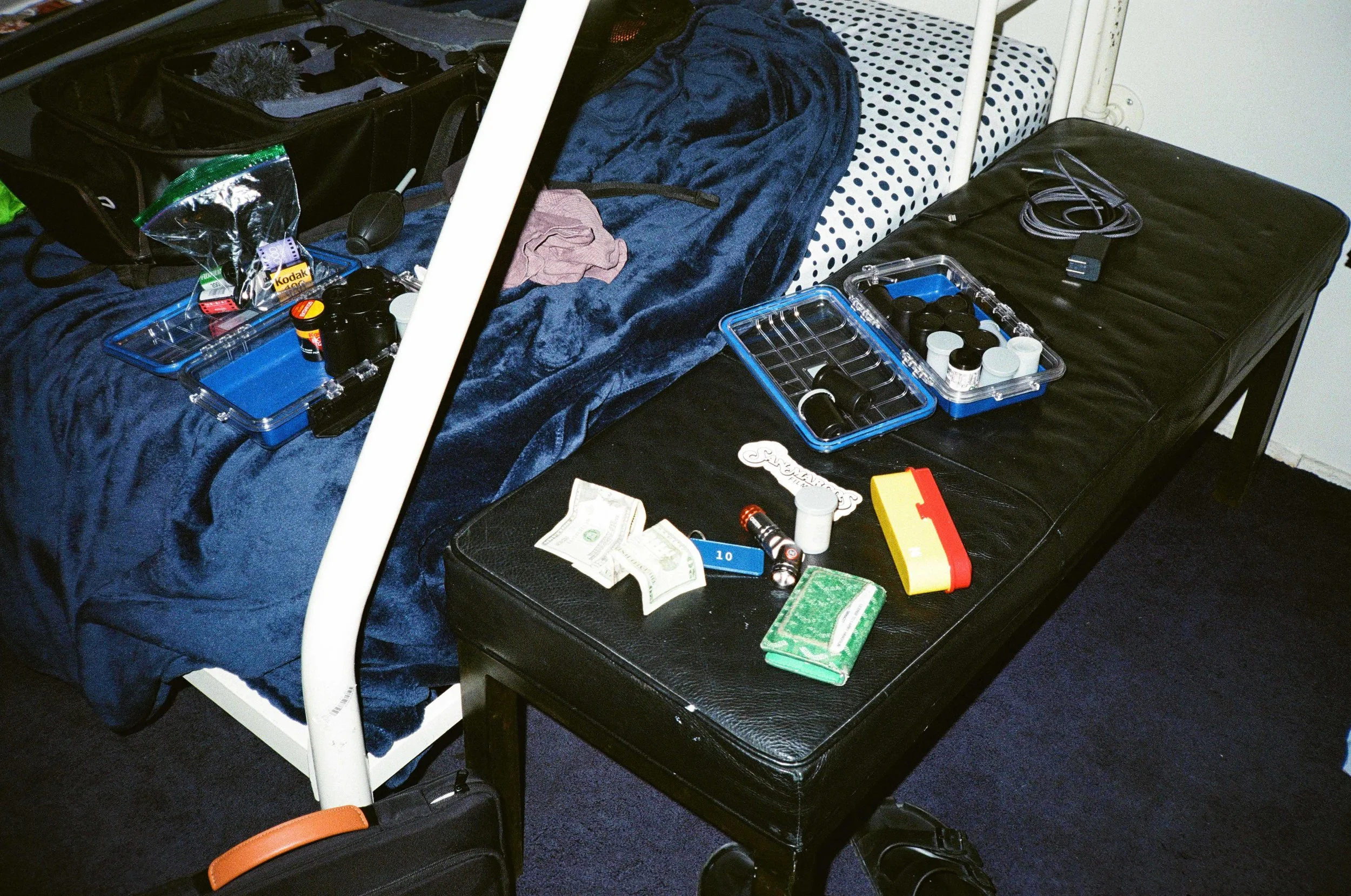 A bed with a blue towel and pink cloth on it, next to a black bag with electronic items, charging cables, and a jar of pills. A black table has a container with pills, a pack of gum, a flashlight, and dollar bills. The scene appears to be a cluttered room or travel setup.