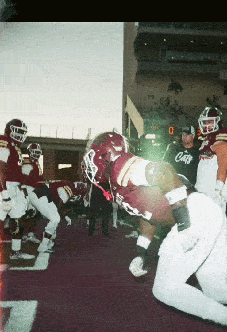 American football team running onto the field with helmets and uniforms, players kneeling and standing near tunnel, stadium entrance in background.