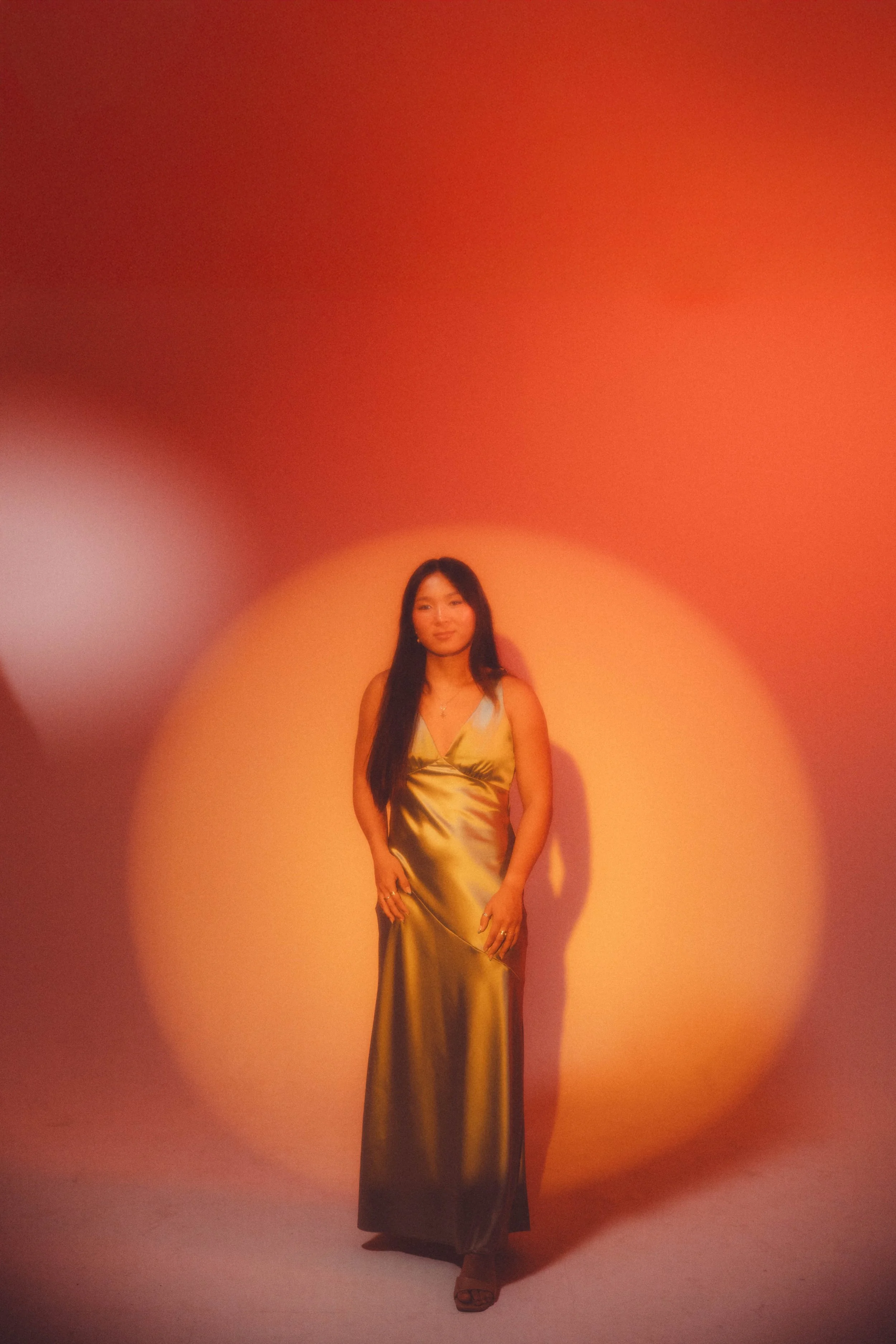 A woman with long dark hair wearing a shiny gold satin dress standing against a warm orange background. The lighting creates a soft gradient effect around her.