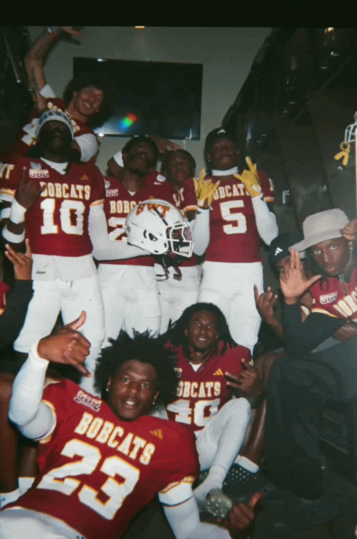A group of football players in red jerseys with 'BOBCATS' and numbers on them, some making hand signs, gathered in a locker room or similar setting.
