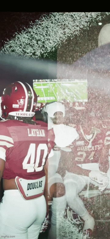 American football players in maroon and white uniforms watching a game on a large screen, with one player focused on the game and standing in front of a soccer field.