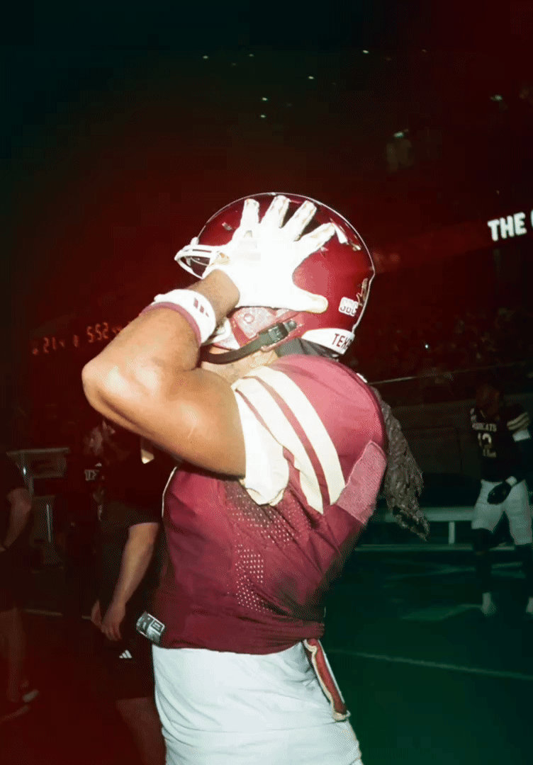 Football player in maroon jersey and helmet adjusting helmet on sidelines of a stadium at night.