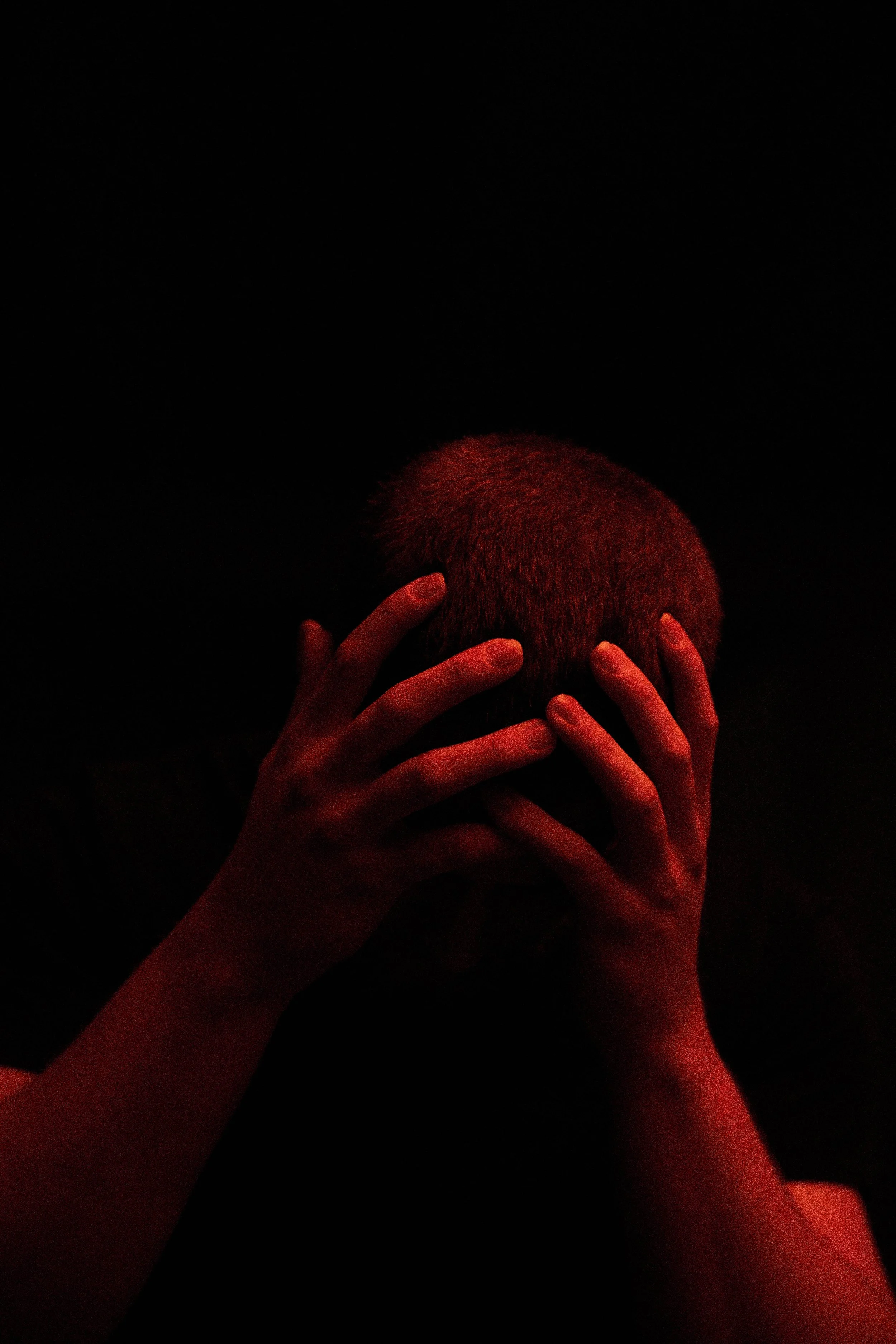 Multiple hands holding a head with short hair against a dark background, illuminated by red light.