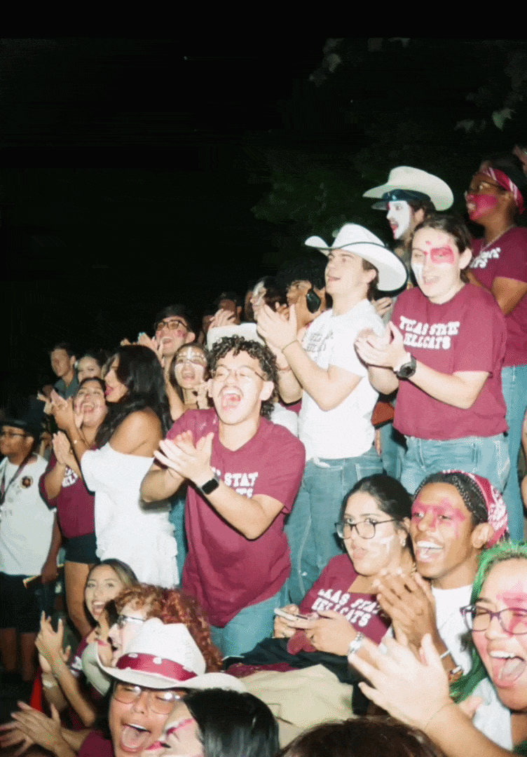 Group of young people at a celebration, some wearing white cowboy hats and face paint, clapping and laughing in a dark outdoor setting.