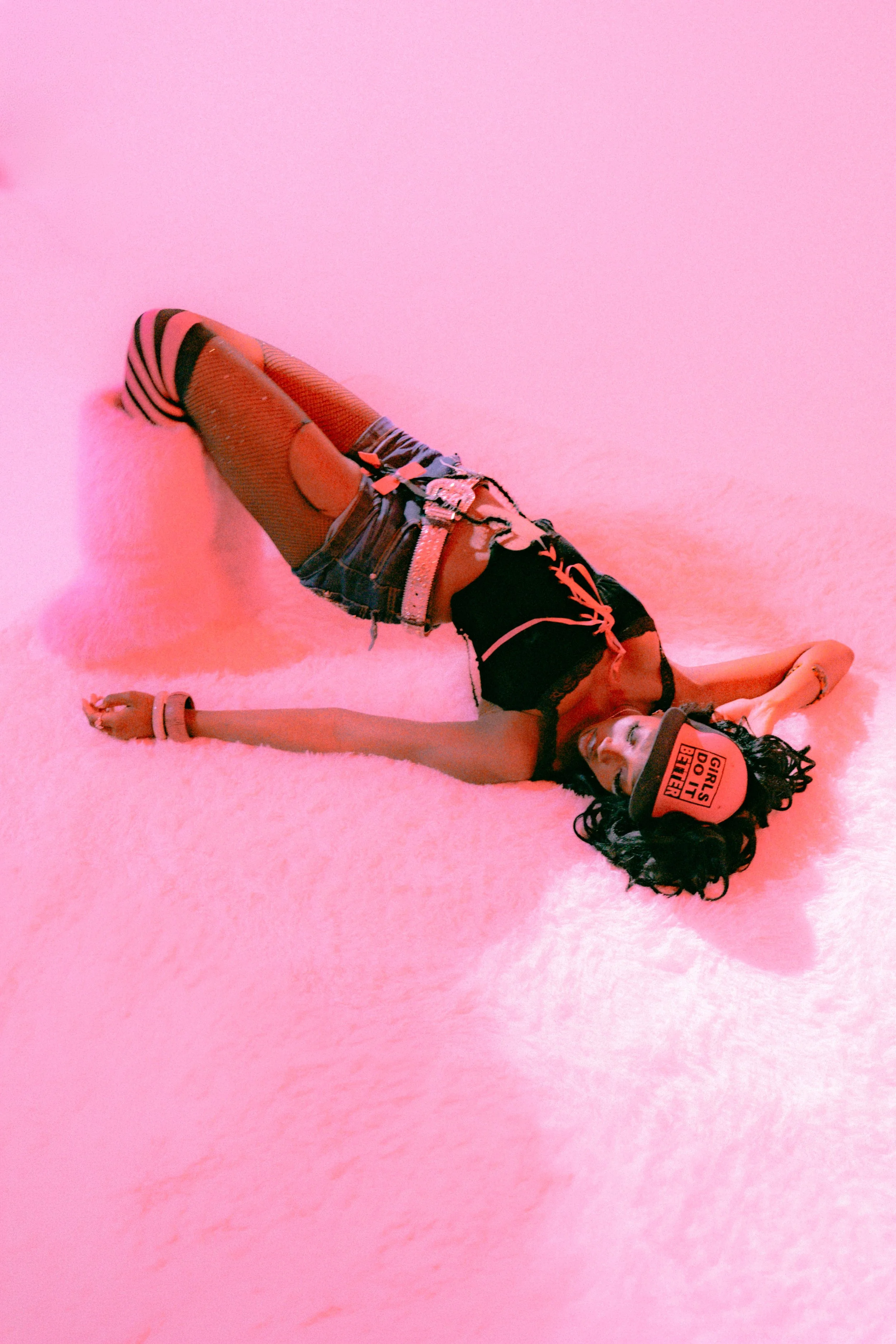 A woman lying on her side on a soft pink surface, wearing a black top with pink lettering, ripped denim shorts, and striped thigh-high stockings, with curly dark hair and a hat that says 'GIRLS DO IT BETTER'.