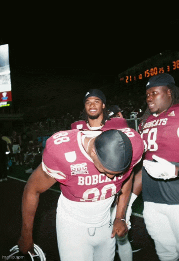 Two football players in maroon and white uniforms on a football field, one bowing his head and the other standing next to him, smiling, during a game or event at night.