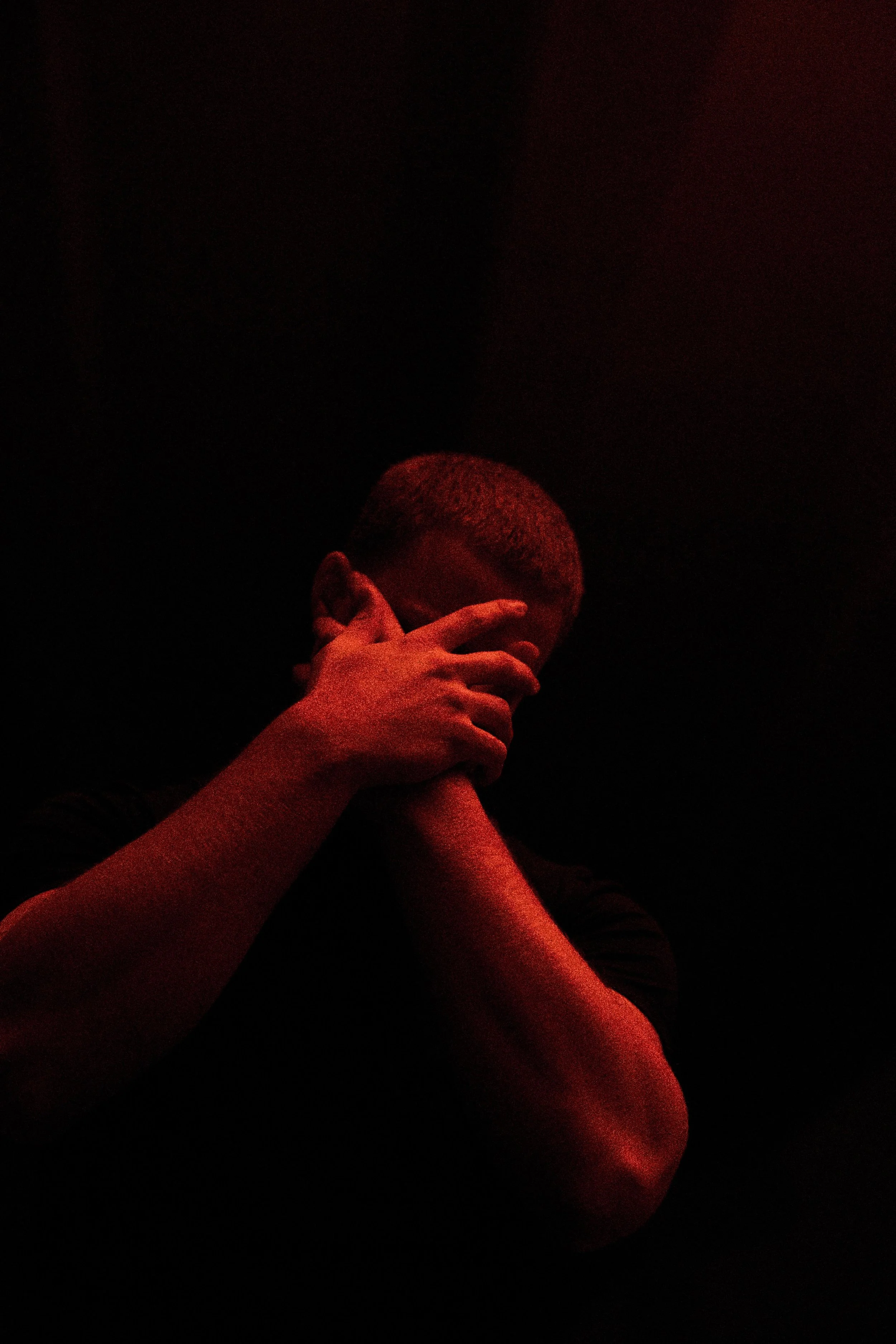 Person with short hair covering face with hand, illuminated by red lighting, dark background.