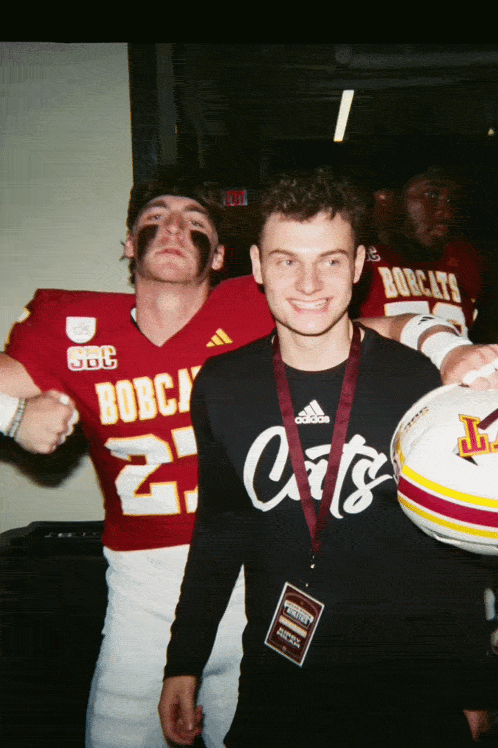 Young man smiling and wearing a lanyard after a football game, with football players in soccer jerseys in the background.