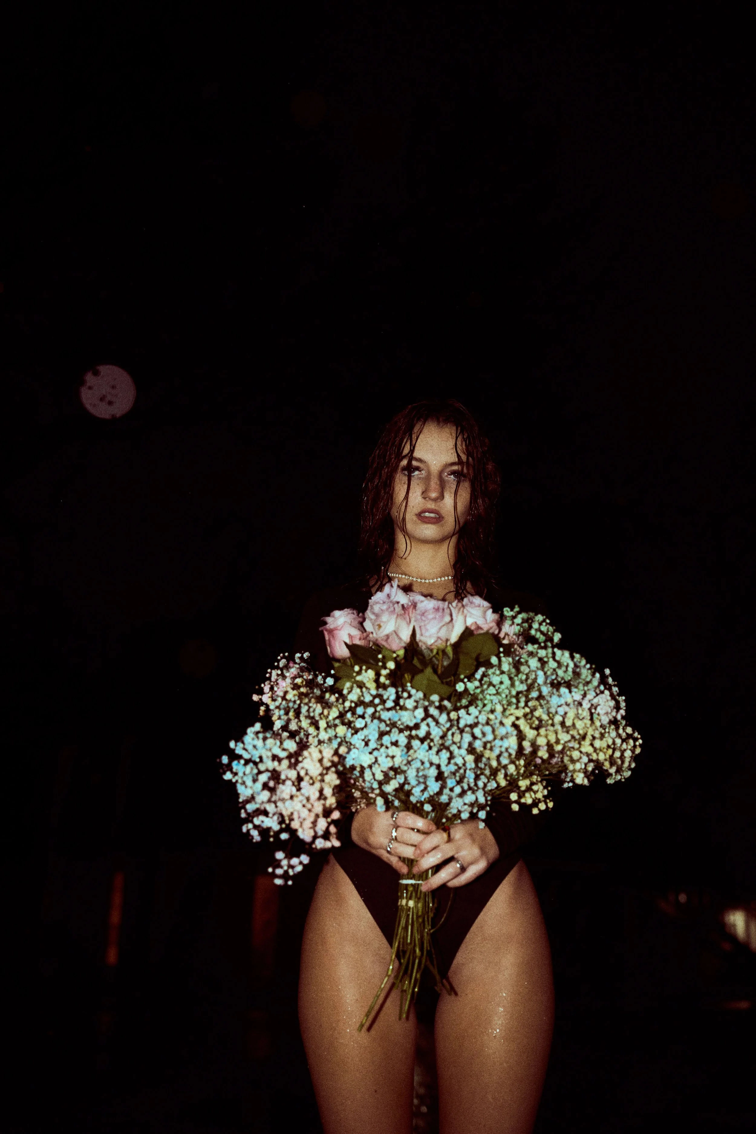 A woman with wet, dark brown hair, wearing black lingerie, holding a large bouquet of pink roses and small white flowers against a dark background at night.