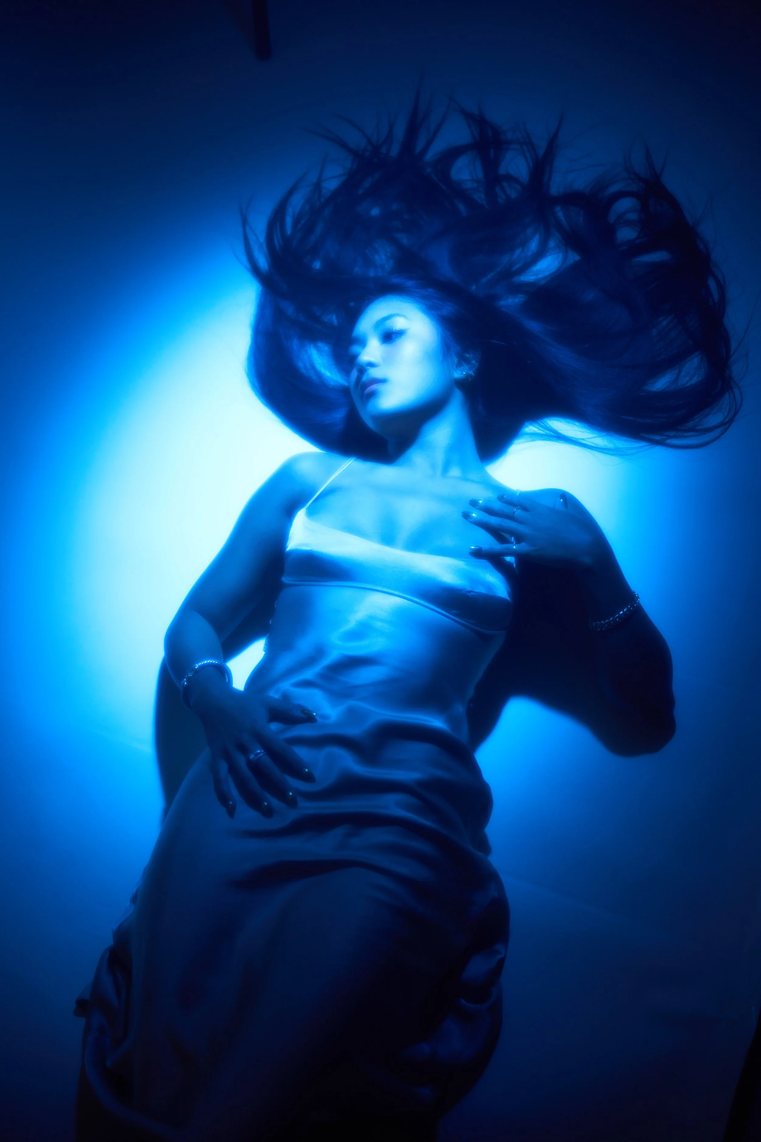A woman with long, flowing hair lying on her back on a blue-lit surface. She is wearing a satin dress and jewelry, with her eyes closed and hand resting on her chest. The lighting creates a glowing, ethereal effect.