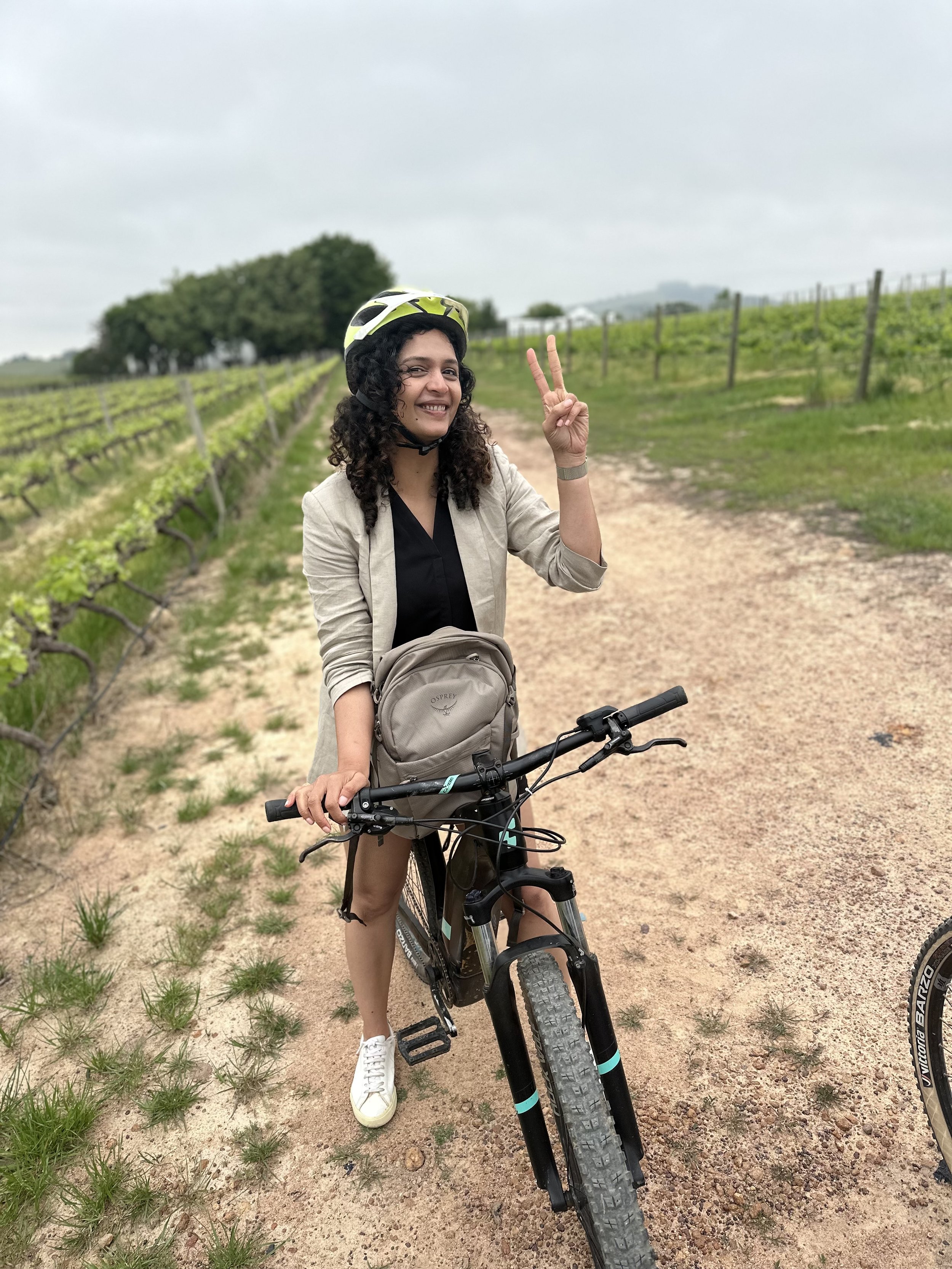Woman with curly hair wearing a beige jacket, black shirt, white sneakers, and a helmet, standing with a mountain bike on a dirt path in a vineyard with green rows of grapevines and trees in the background, on an overcast day.
